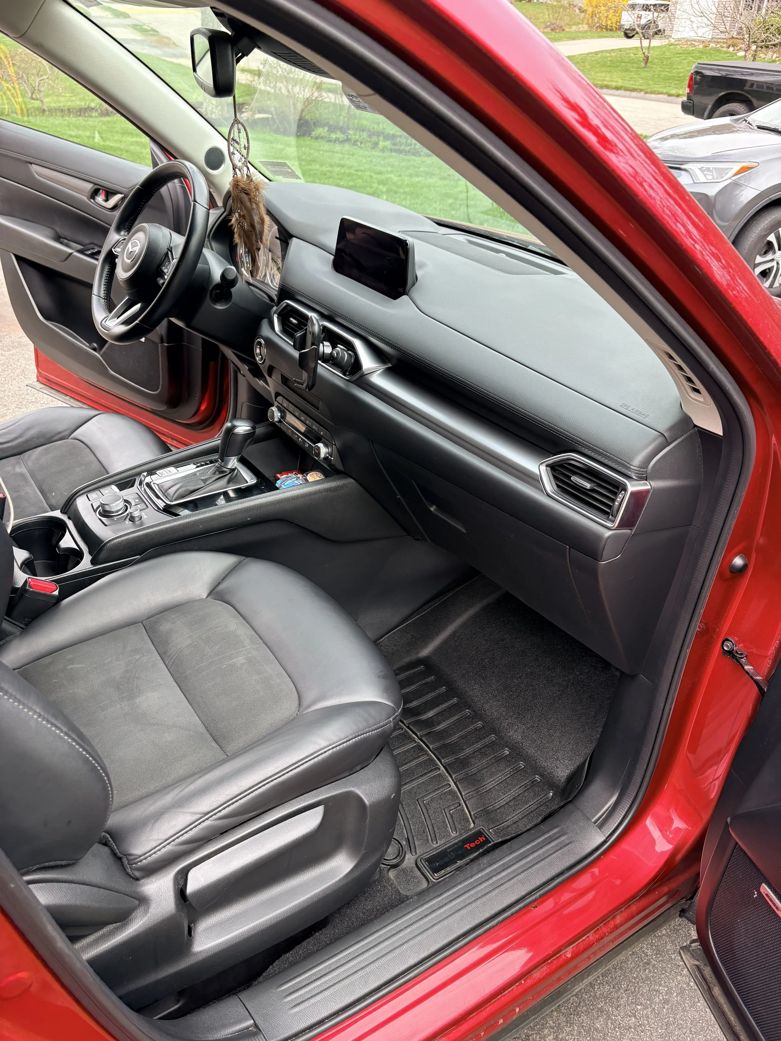 Inside of a red car shows a black leather driver's seat, dashboard, steering wheel with a Mercedes-Benz emblem, and a touch screen display. The car is parked outside on a driveway with green grass and other vehicles visible through the windows.