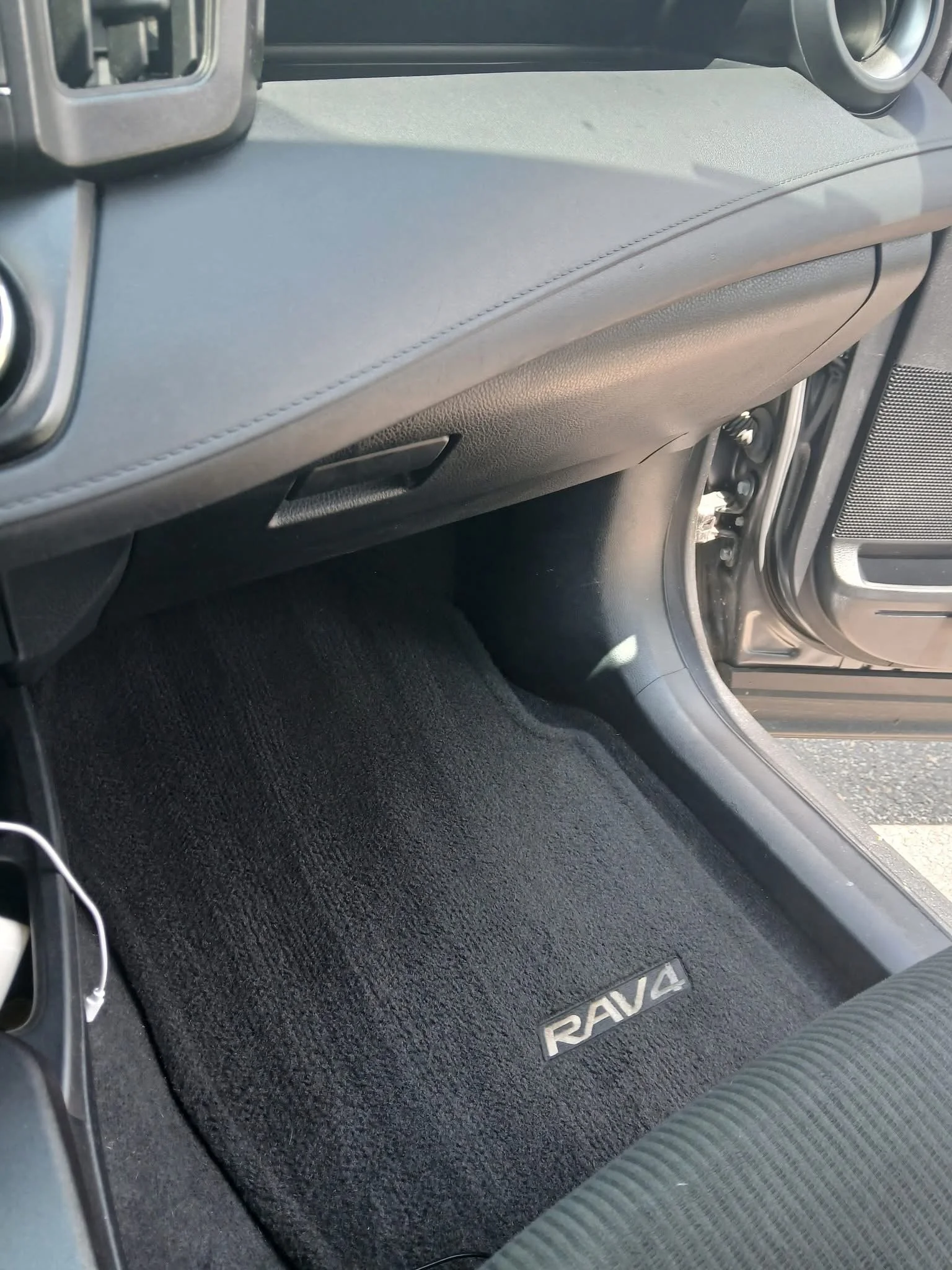 Interior view of a car showing the black carpeted floor mat with 'RAV4' branding, part of the dashboard, and a portion of the driver's seat.