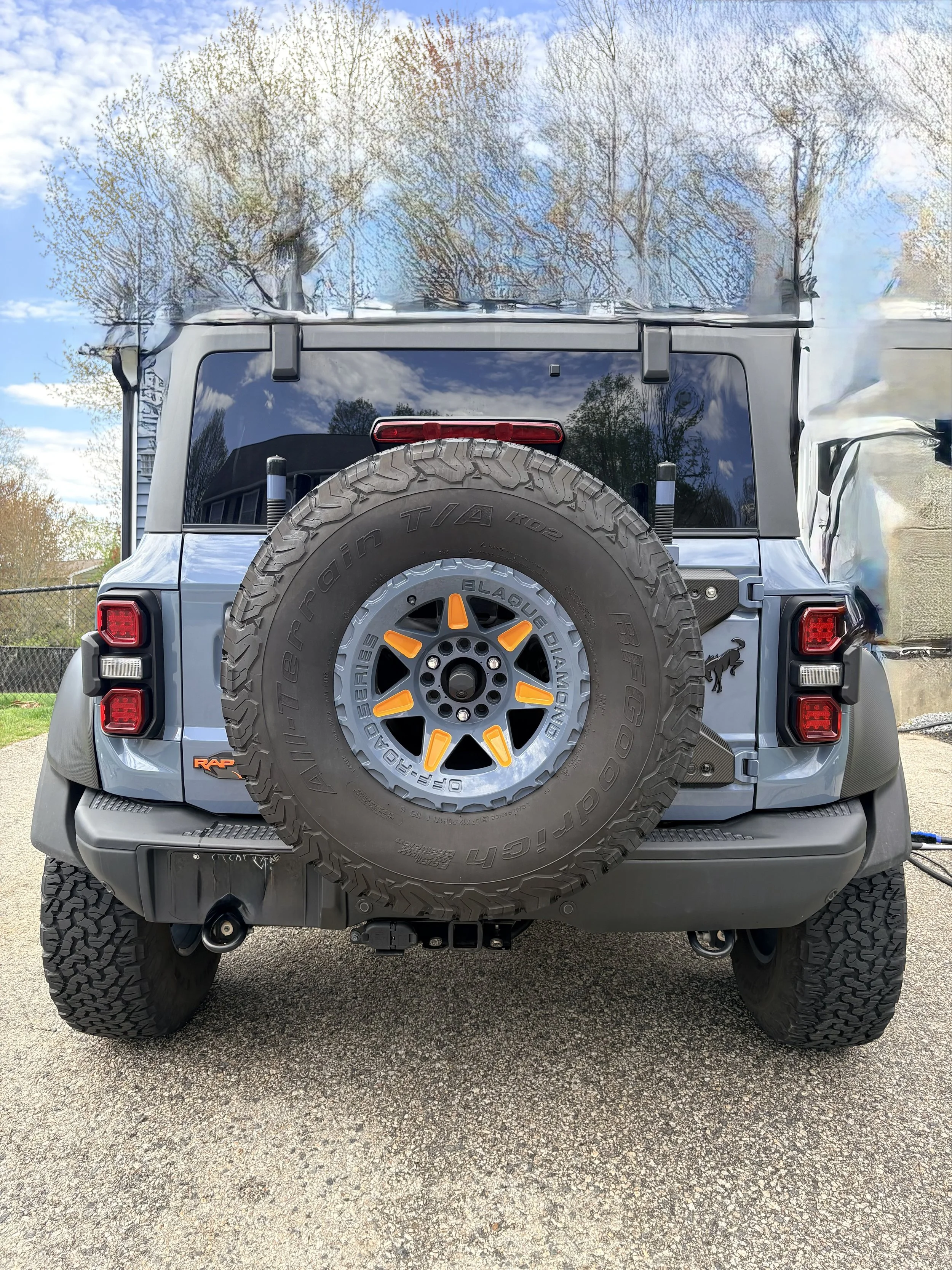 The rear view of a blue Jeep SUV with a mounted spare tire on the back, parked outdoors on a paved surface, with trees and a blue sky with clouds reflected on the vehicle's rear window.