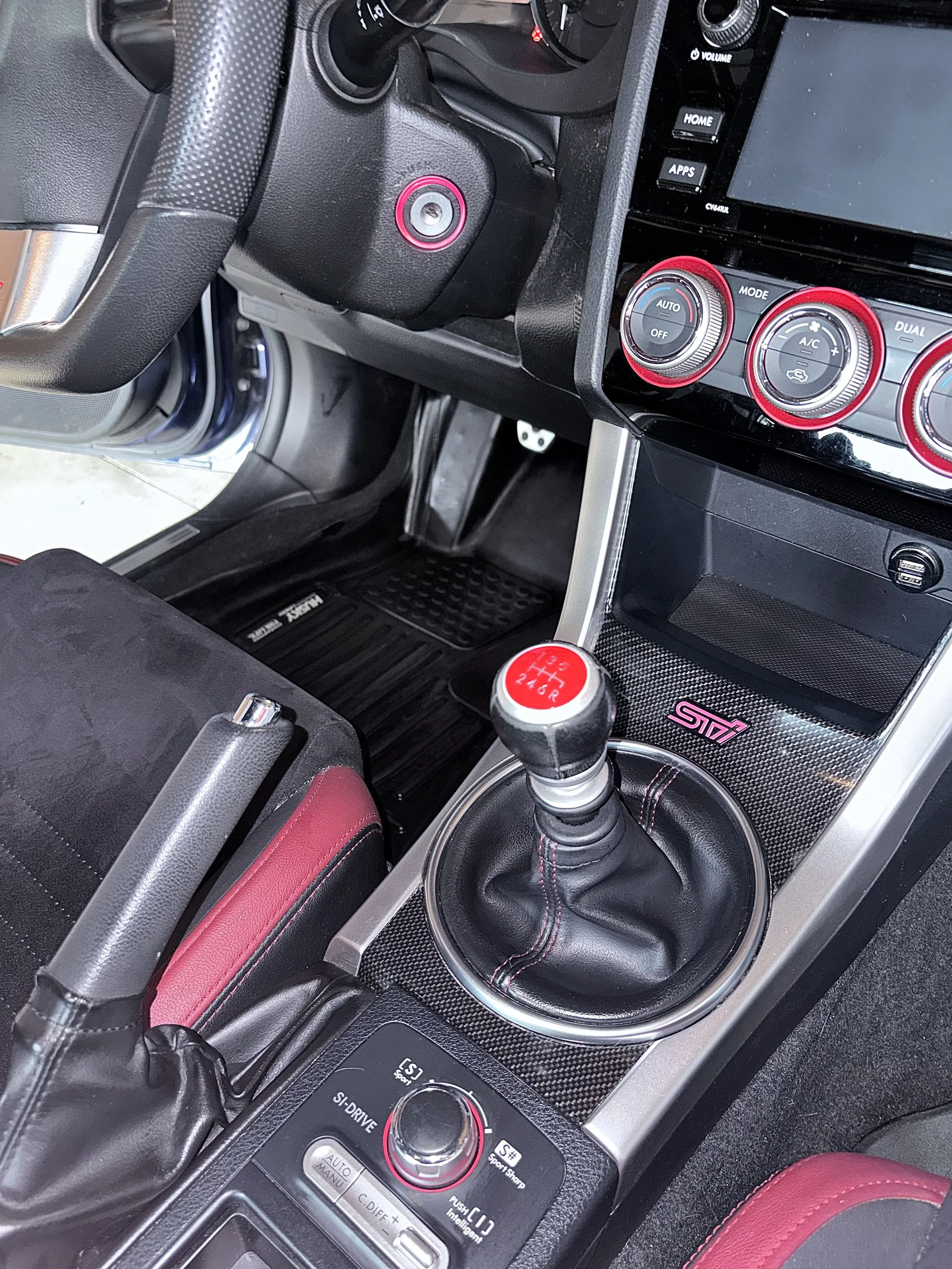 The interior of a car showing the center console with a gear shift, climate controls, and a display screen. The gear shift has a red and white button, and the surroundings feature various buttons and dials with red accents.