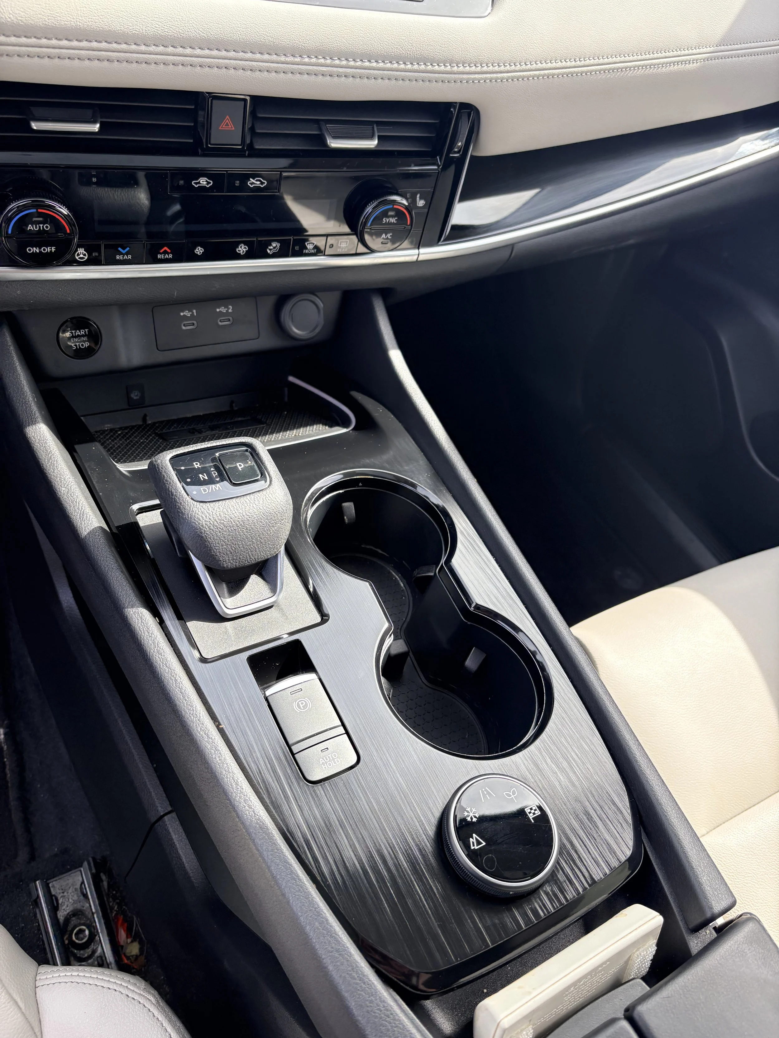 The image shows the center console of a modern vehicle, featuring climate controls, a gear shift, a cup holder, and various buttons and dial controls.