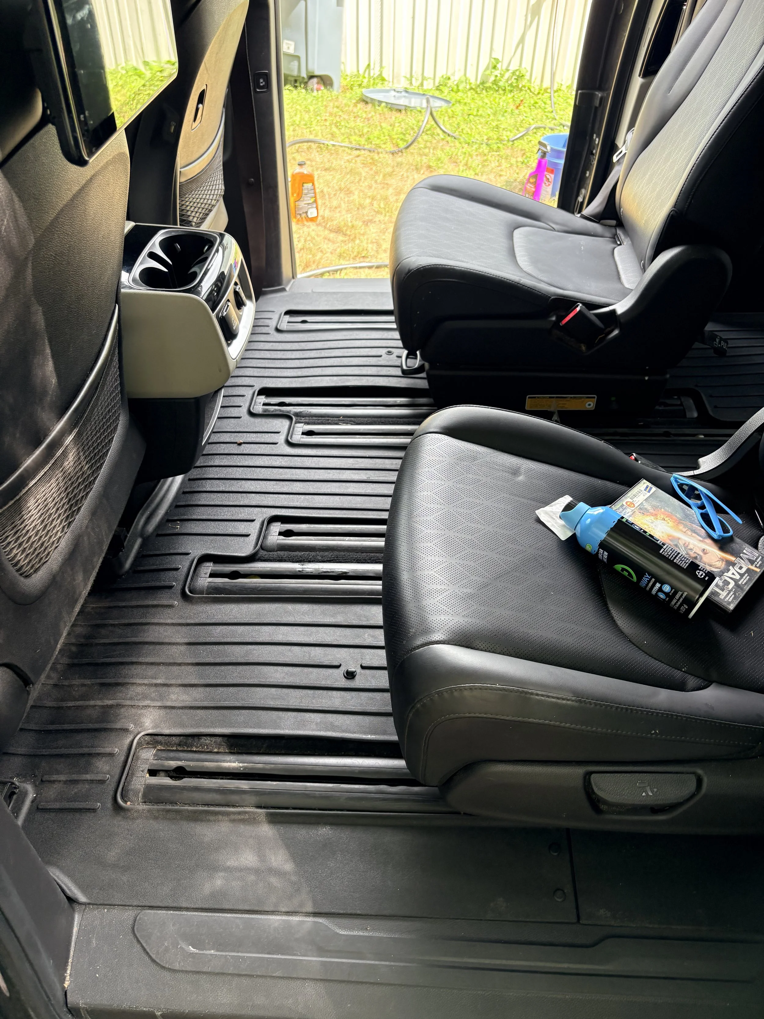 The front part of a vehicle's interior showing two black seats, a center console with cup holders, and a view of the outdoors with grass and a fence.