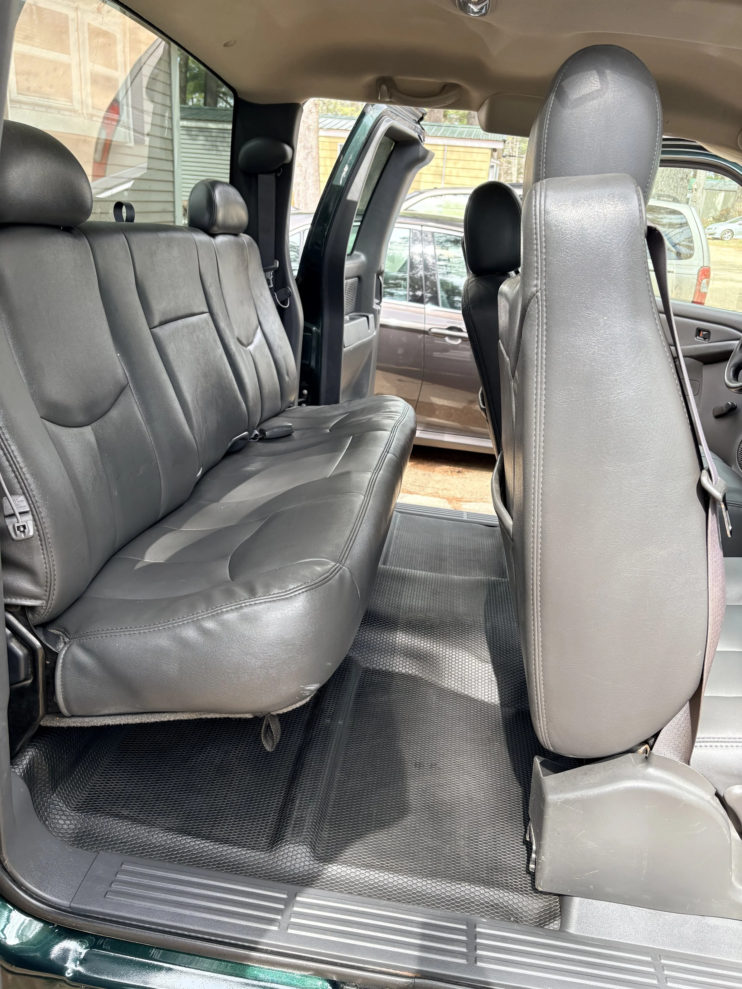 The interior of a vehicle showing the back seats and front seats with all seats covered in black leather. The vehicle has a black rubber floor mat and an open door revealing outside cars and a building.