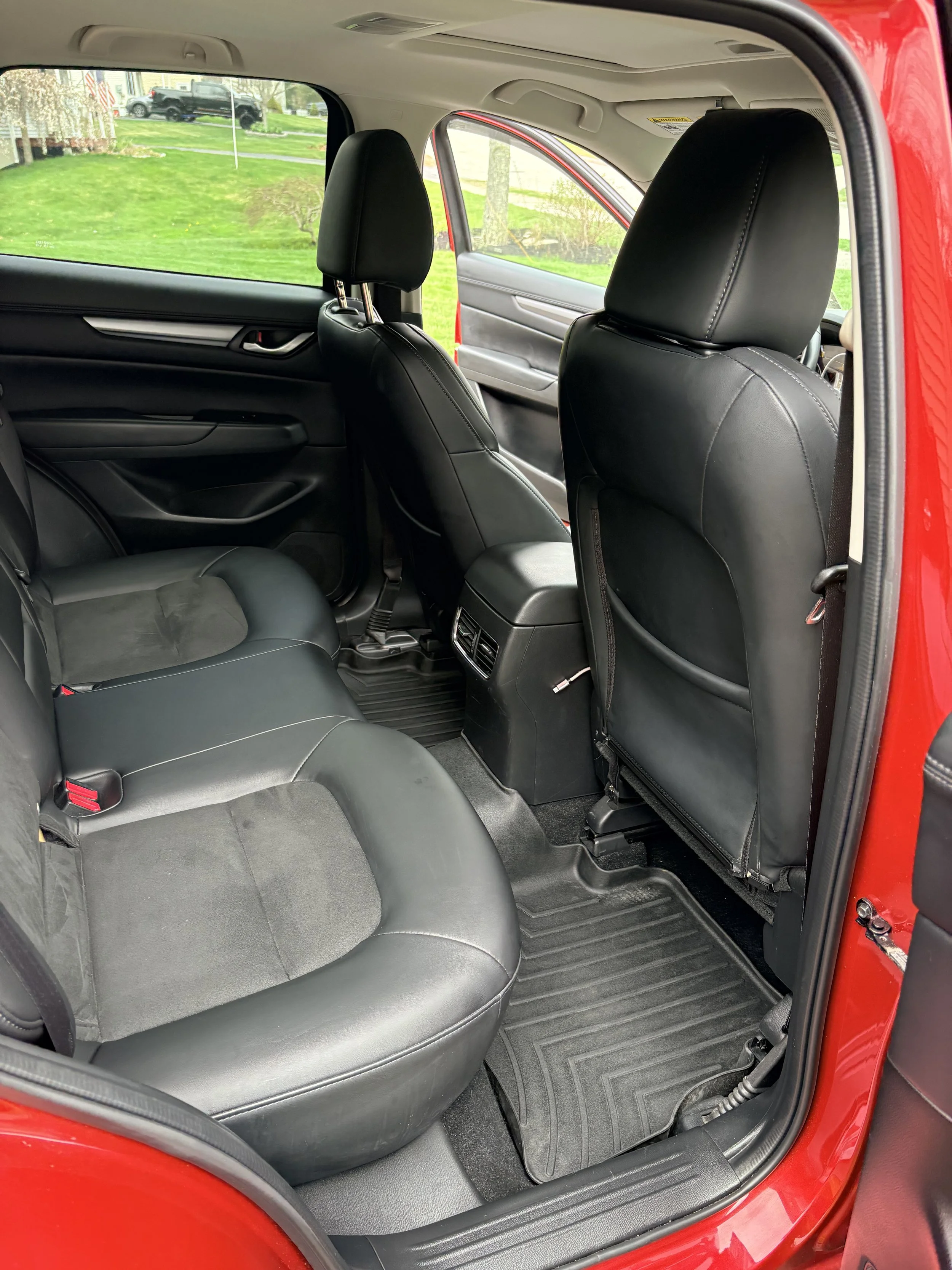 The interior of a red vehicle showing black leather seats, with the rear passenger seat and front driver's seat visible. The door is open, revealing the window and a background of a green lawn and trees.