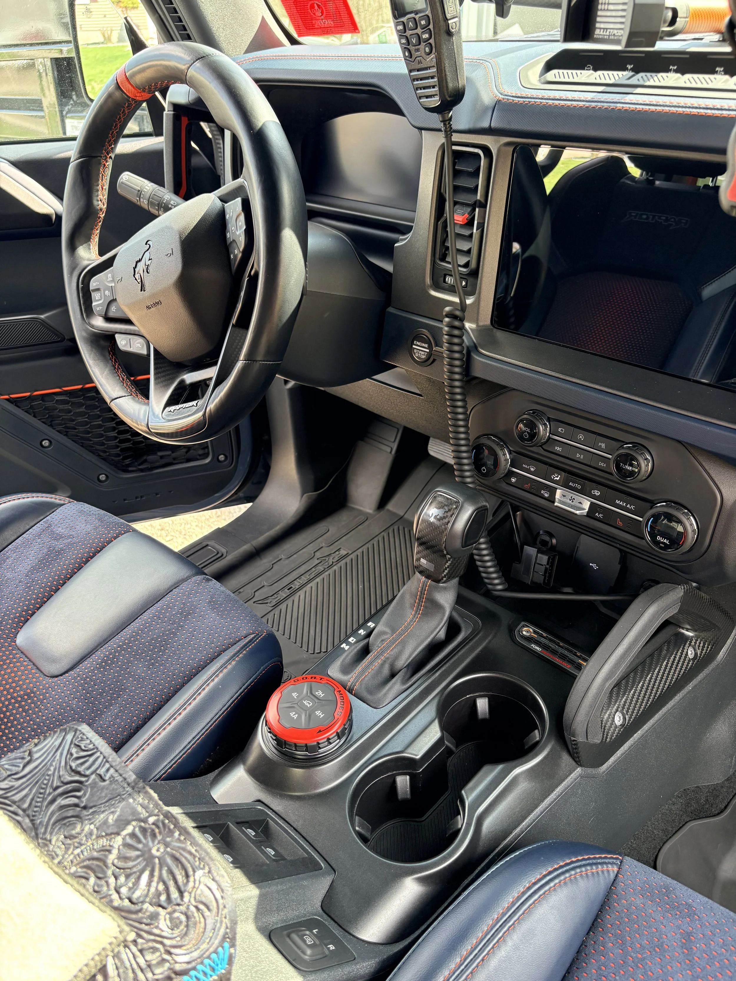 Interior of a modern vehicle featuring a steering wheel with orange stitching, digital display screen, control buttons, a radio system, climate controls, a gear shift with a red rotary dial, cup holders, and racing-style bucket seats with red stitchi