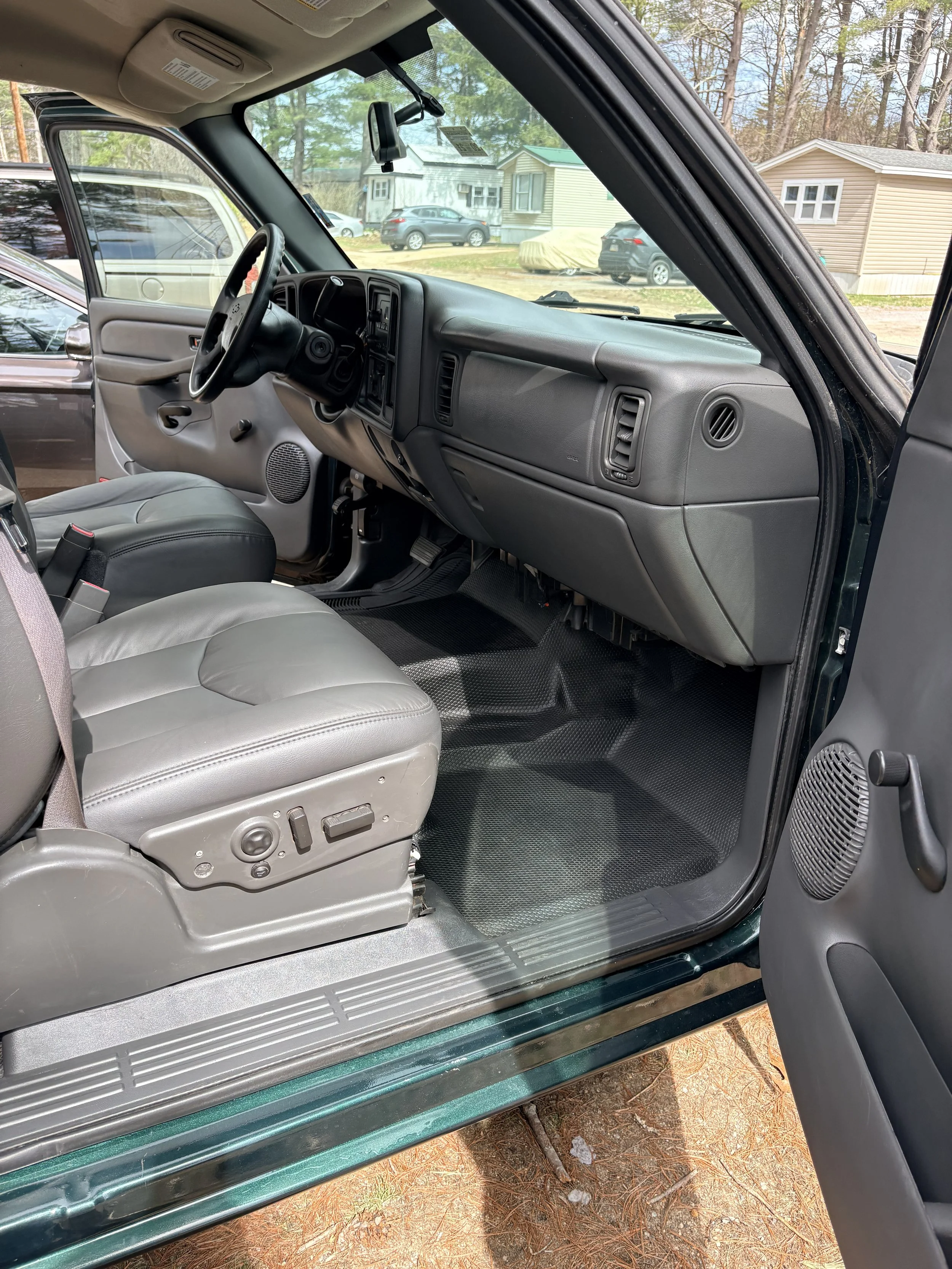Interior view of a vehicle with gray seats and dashboard, viewed from the open front passenger door, showing a rural outdoor scene with trees, houses, and parked cars.
