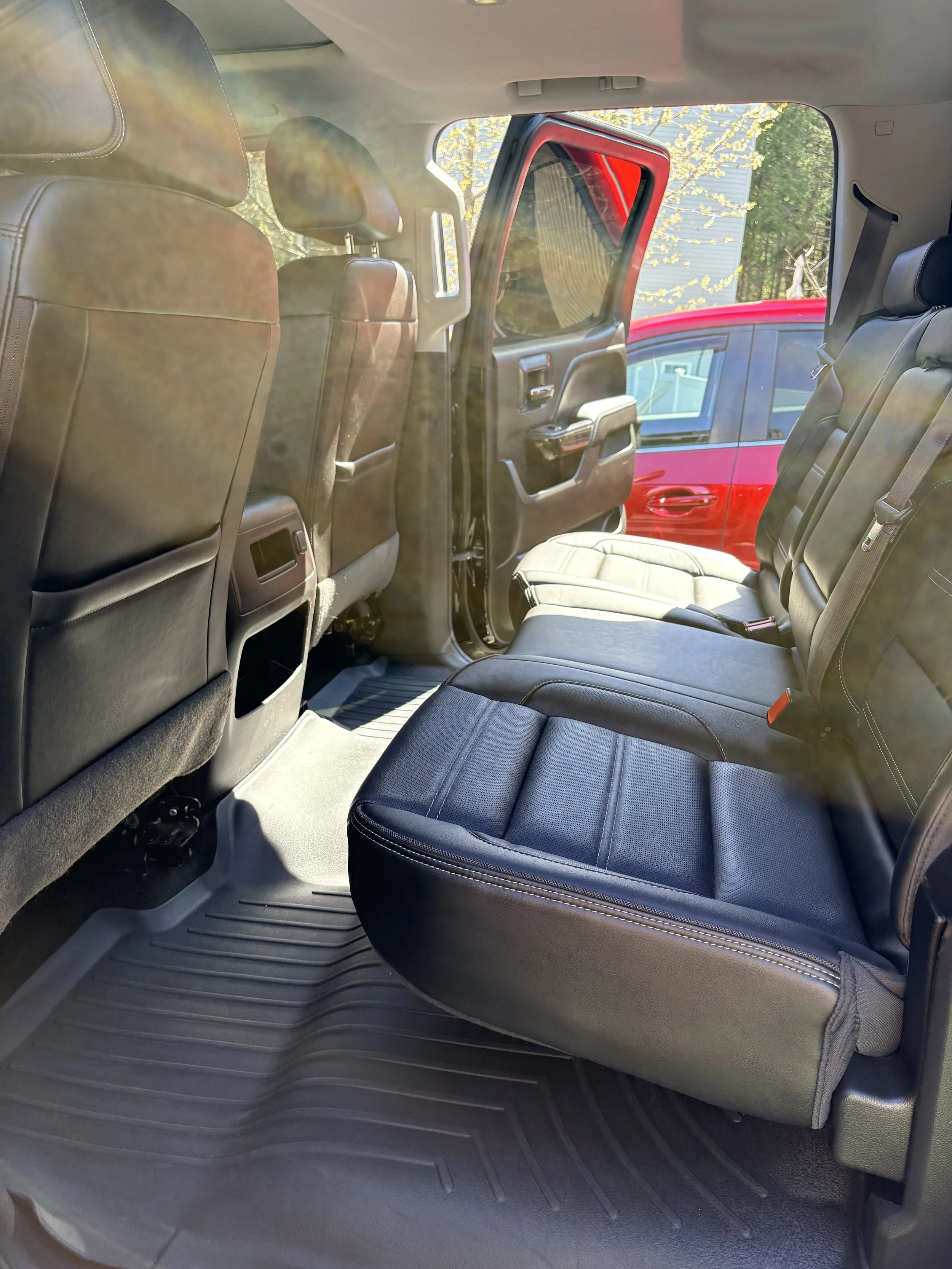 Interior of a vehicle showing the backseat with black leather seats, a rubber floor mat, and the back of the front seats. Outside, another red vehicle and some trees are visible through the windows.