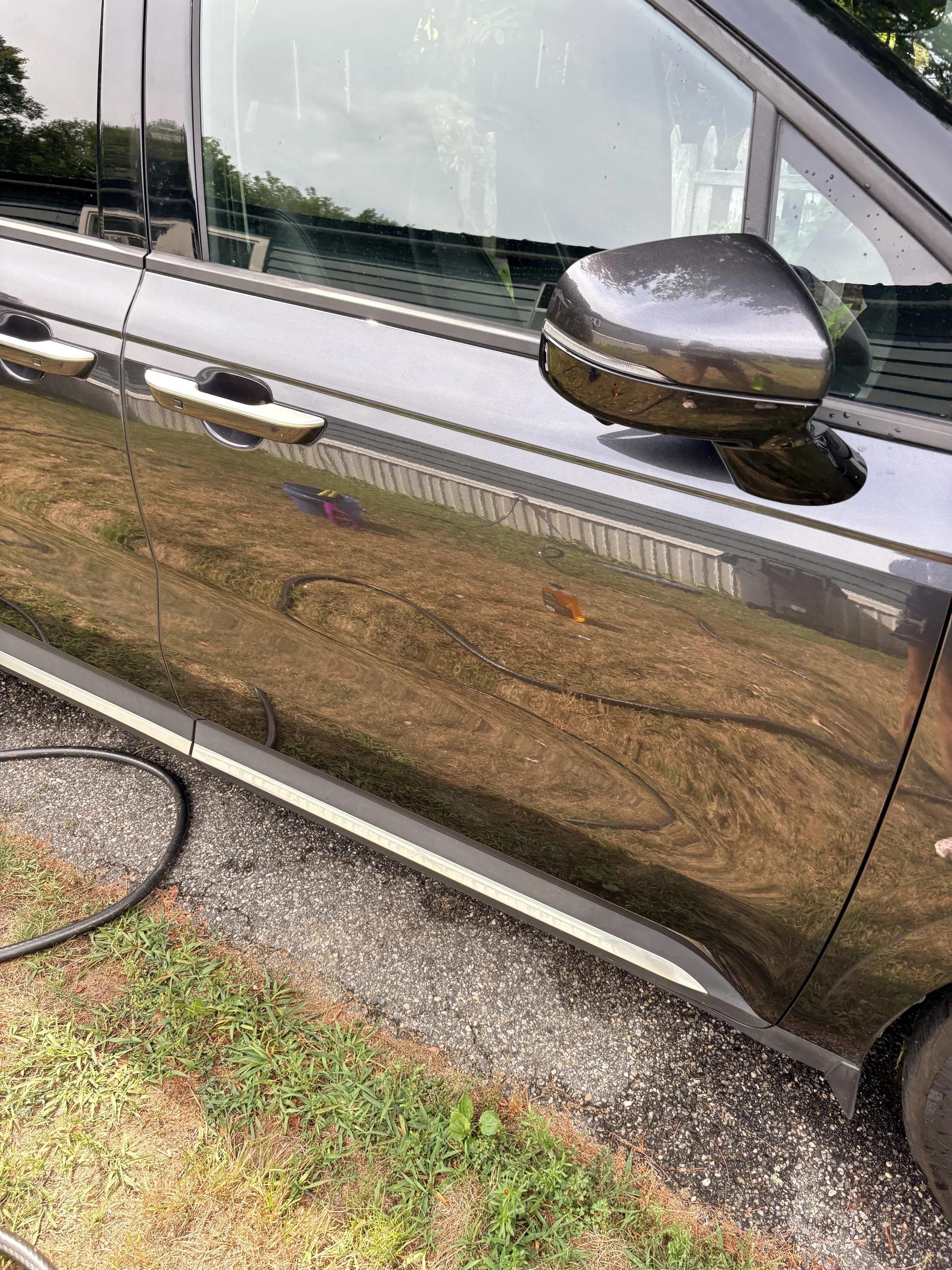 Side view of a silver or gray vehicle with a side mirror, parked on a gravel and grassy area.