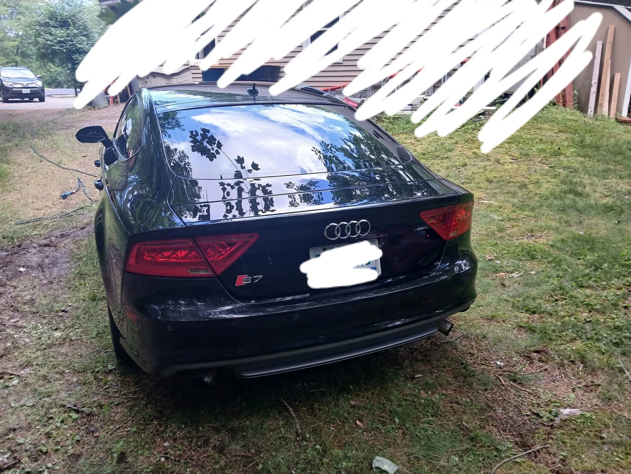 A black Audi S7 car parked on grass in front of a wooden house with a brown fence, with trees and a car in the background and a sky reflected in the car's roof.