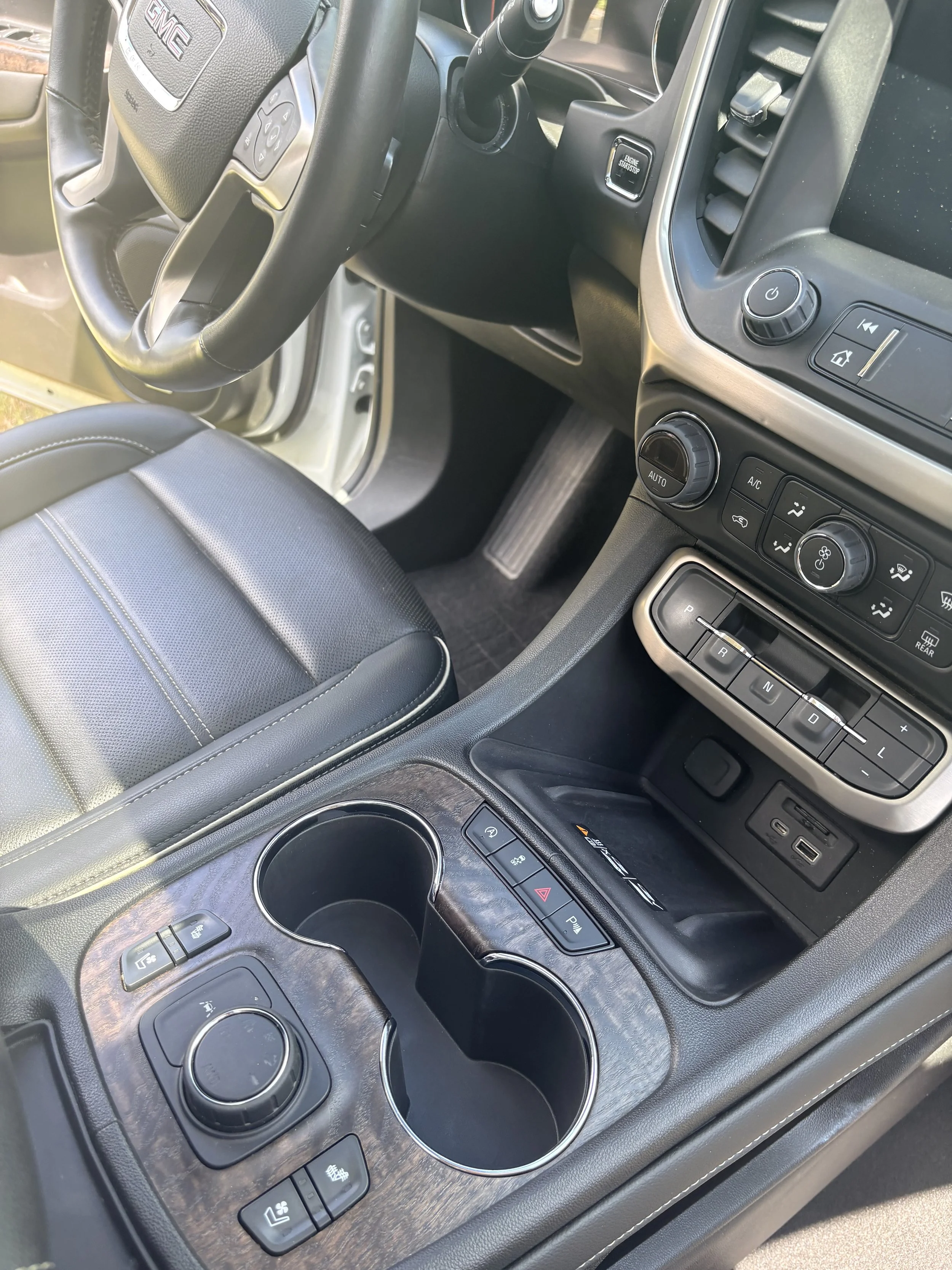 Interior view of a vehicle dashboard showing steering wheel with control buttons, climate controls, gear shift, and cup holders.