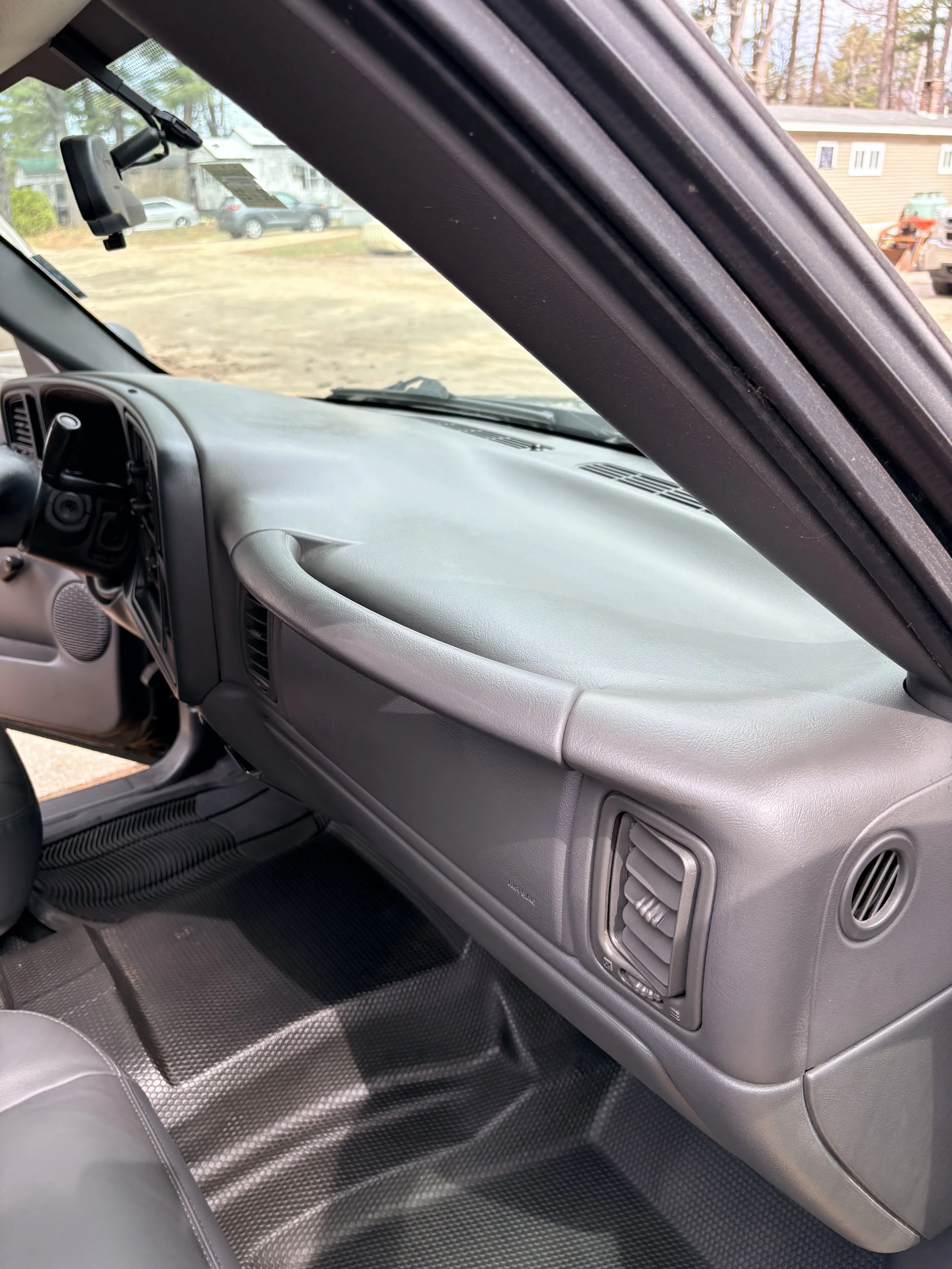 The interior front passenger side of a vehicle, showing a gray dashboard, air vent, glove compartment, and a black rubber floor mat with circular patterns.
