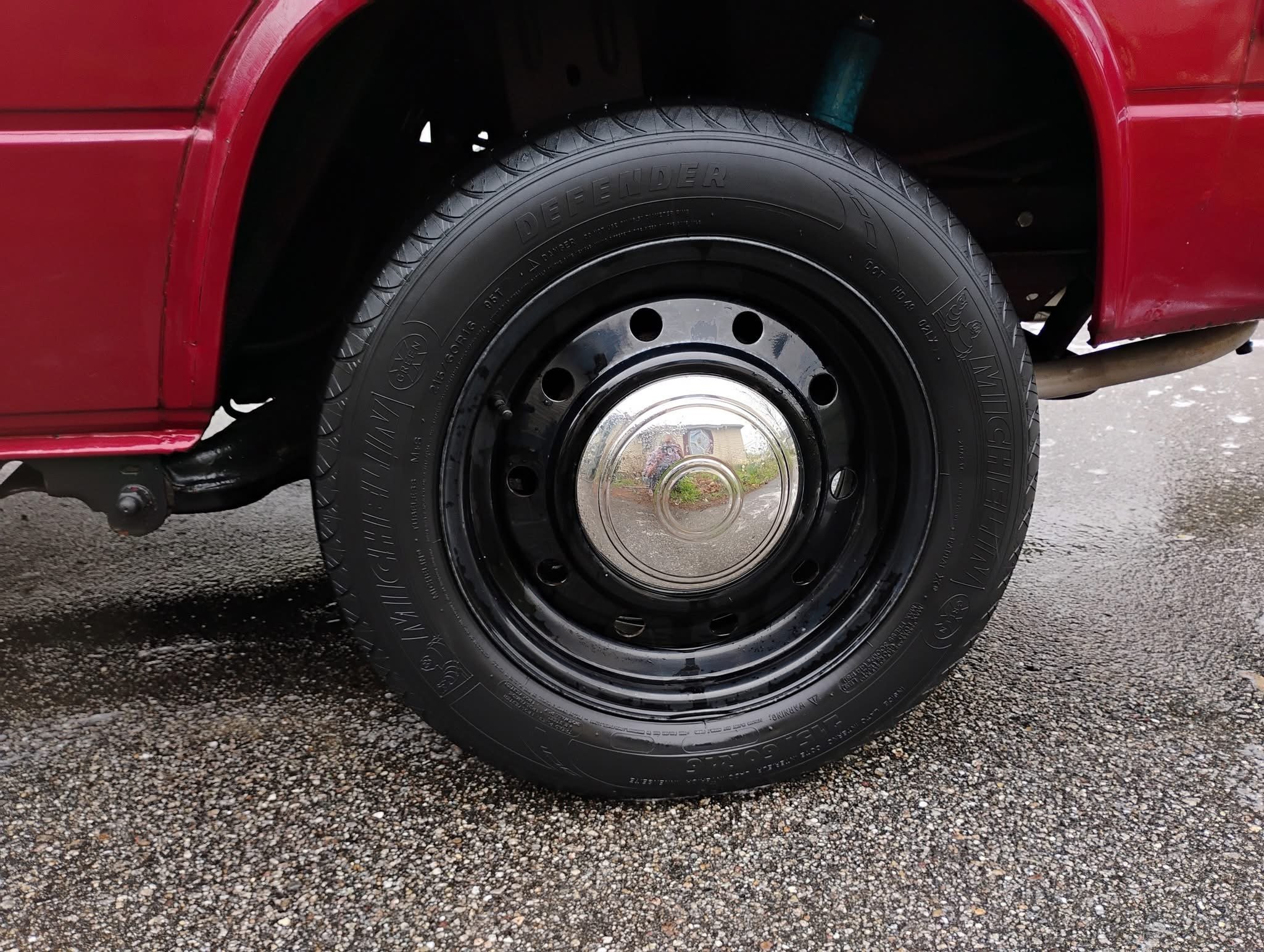 Close-up of a vehicle's front black tire with a shiny hubcap, set on a gravel surface with red bodywork visible on the side.