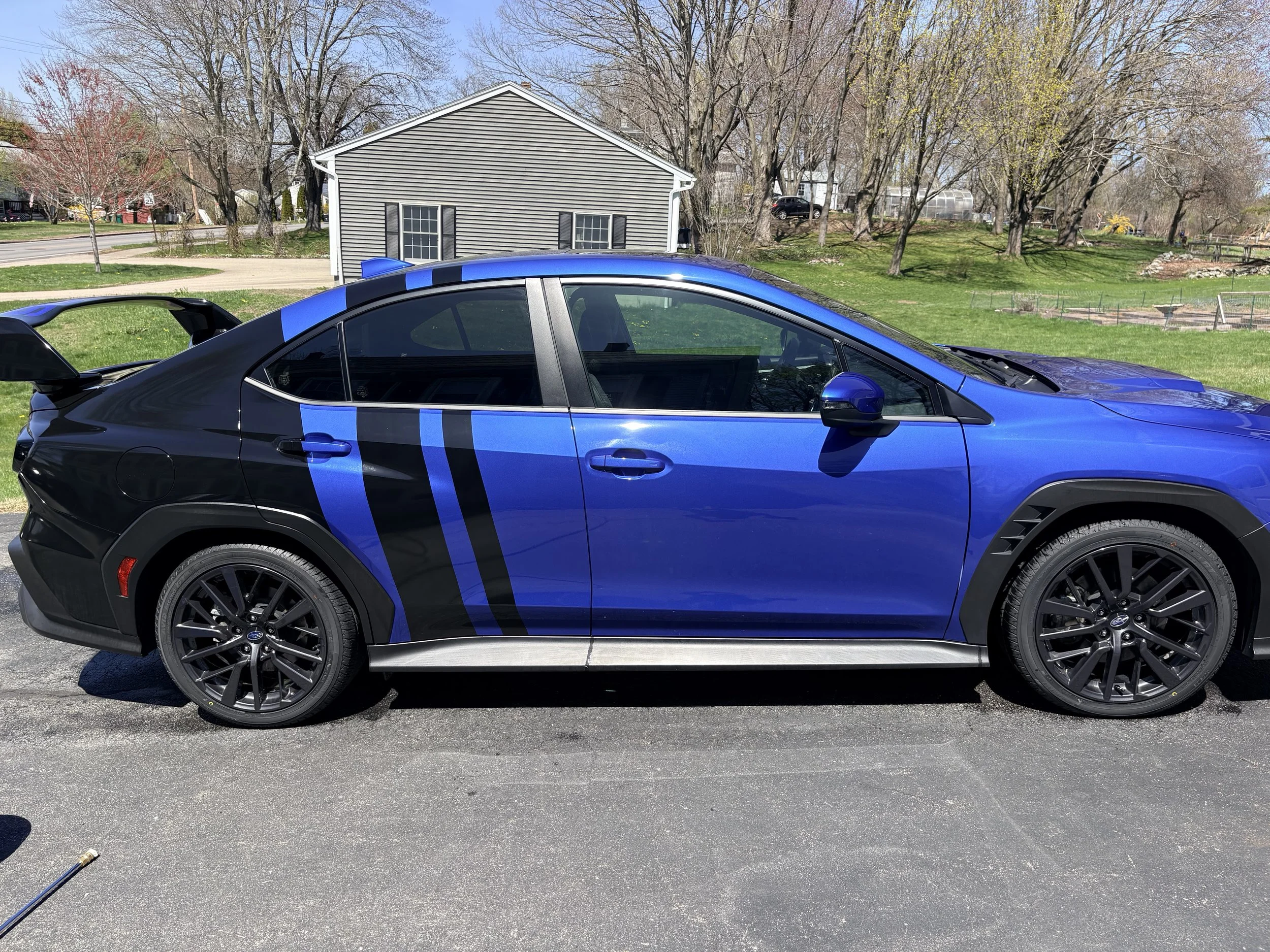 A blue and black sports car parked on a paved driveway in front of a house with gray siding, surrounded by leafless trees and grass.