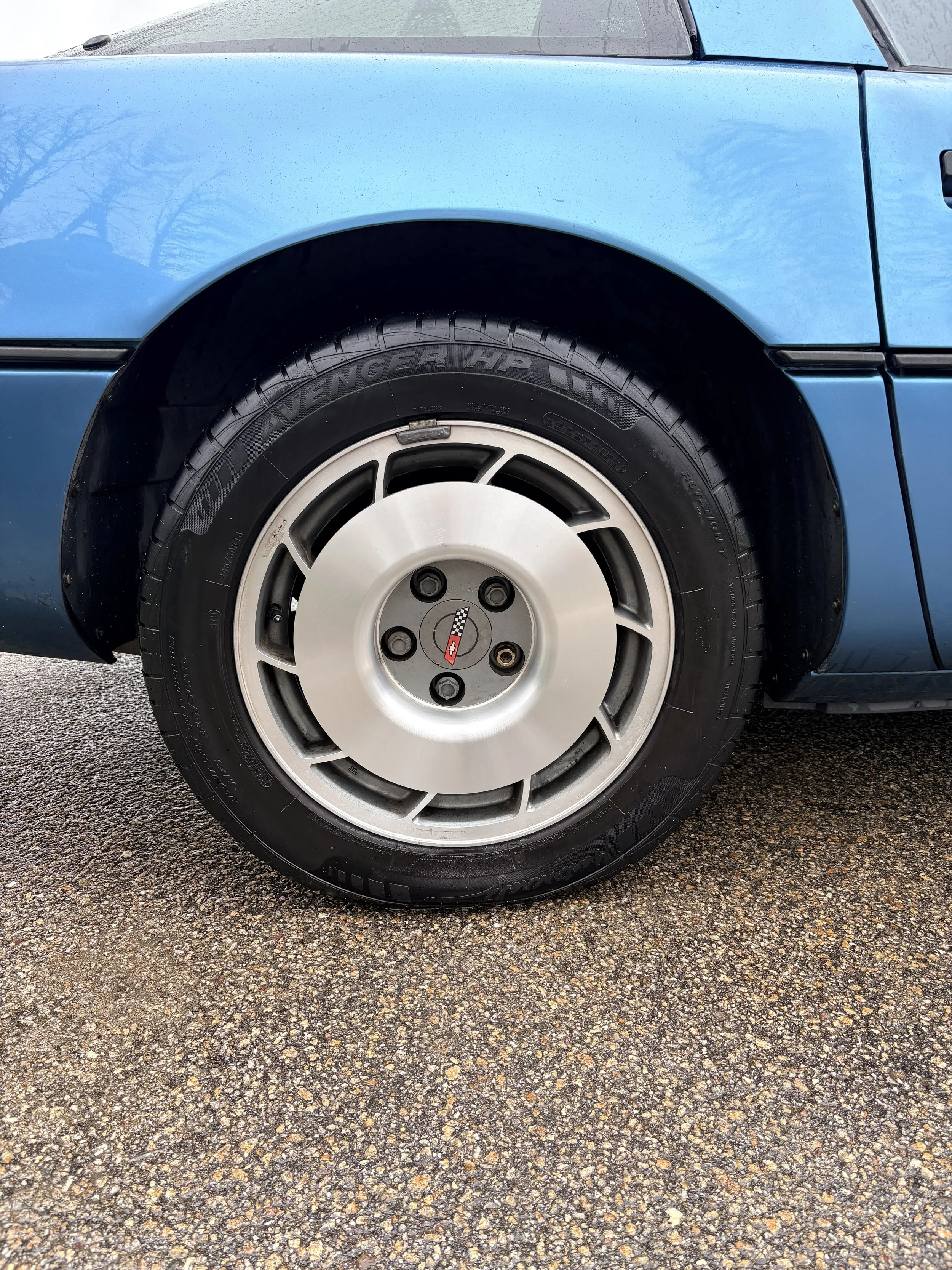 A close-up of the front wheel of a light blue vintage car parked on a gravel surface, showing the tire, wheel, and part of the car's body.