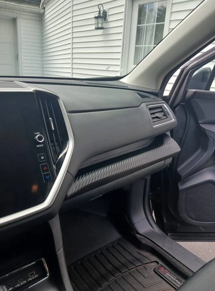 Front passenger side of a modern vehicle interior with a dashboard, air vent, and glove compartment, with an outside house visible through the window.