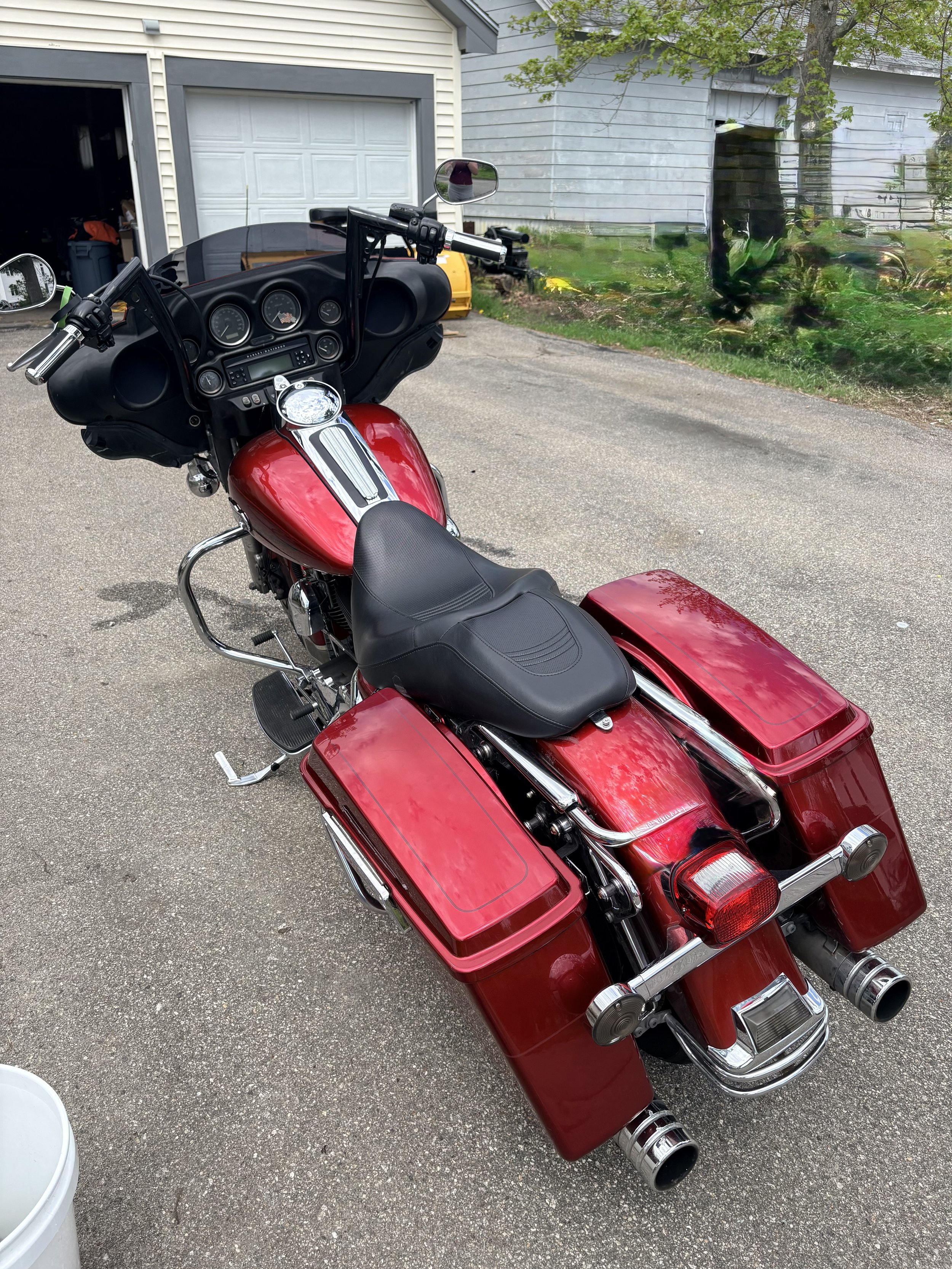 Rear view of a red touring motorcycle with attached saddlebags parked on a driveway in front of a garage and a light-colored house with trees in the yard.