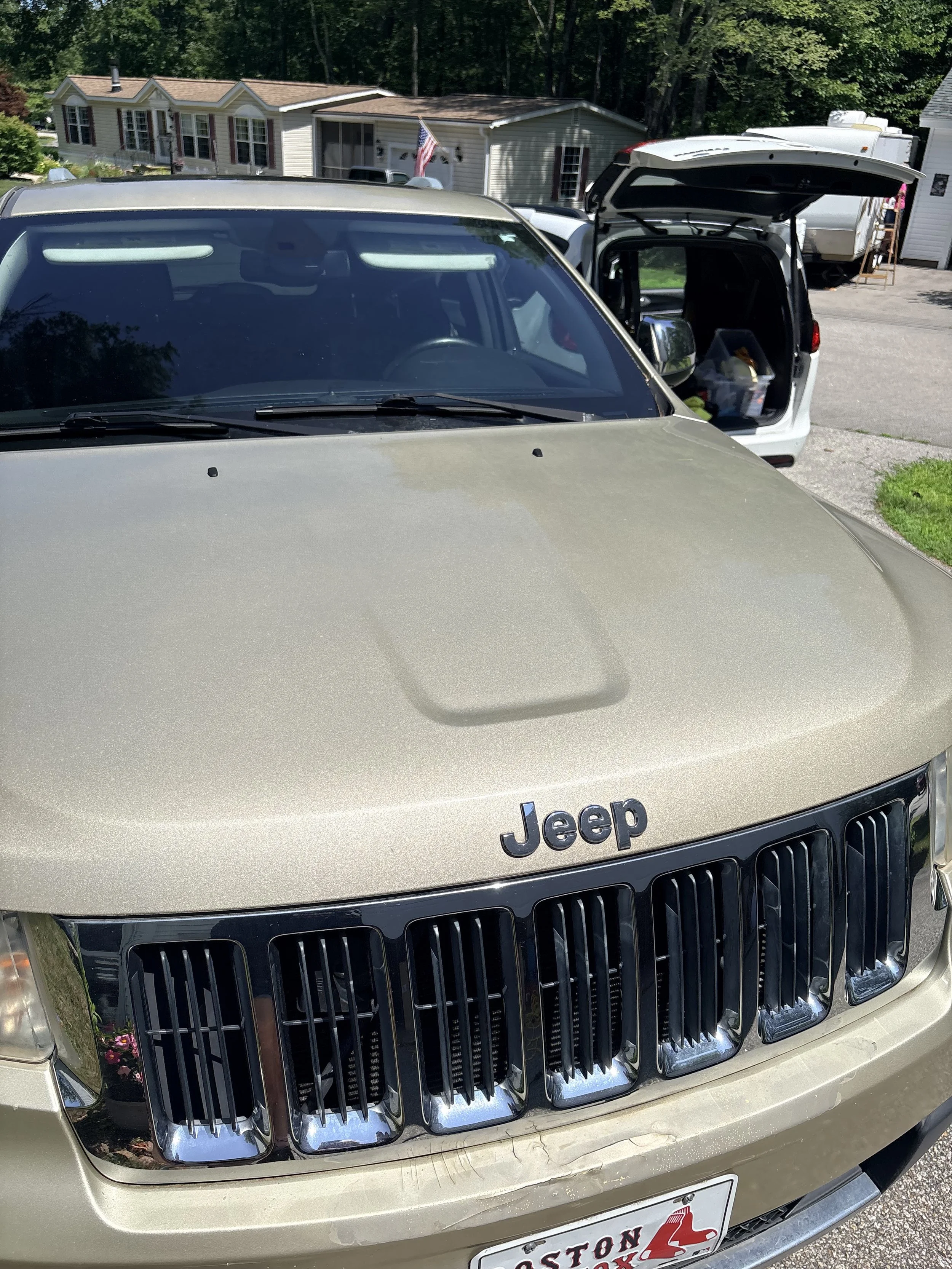 Front view of a beige Jeep vehicle with the hood and grille visible, parked outdoors in a residential area with houses and trees in the background.