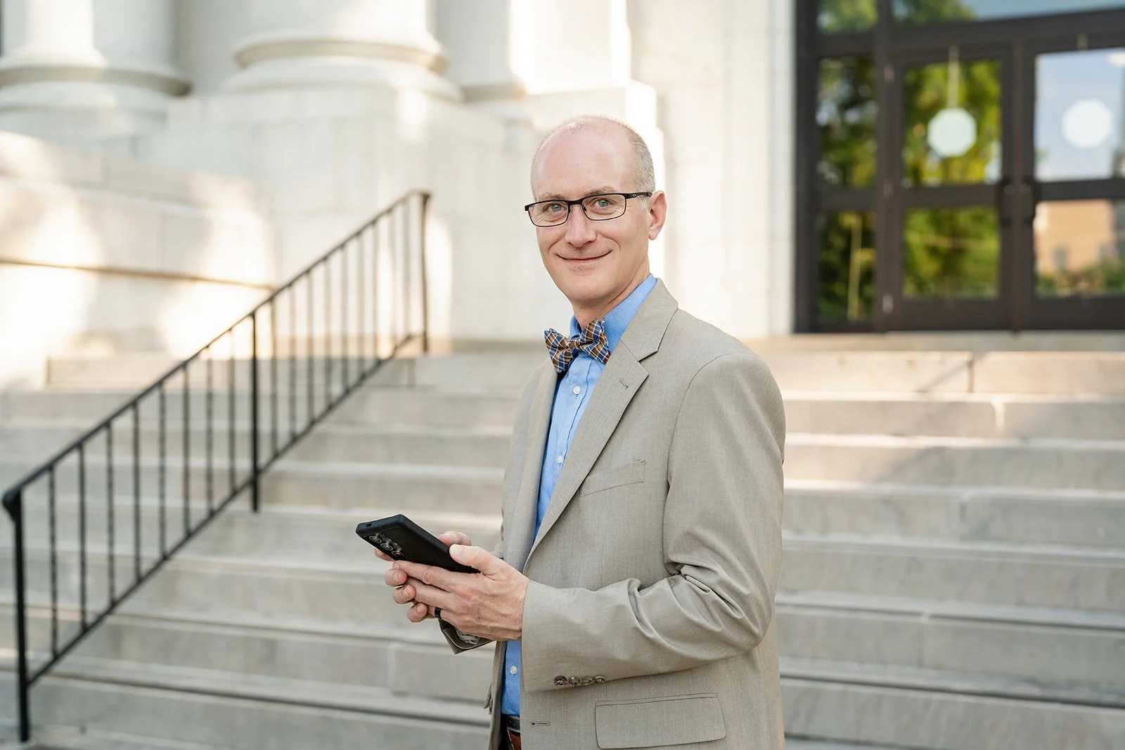 A man in a beige suit, light blue shirt, and bow tie standing on steps outside a building, holding a smartphone and smiling.