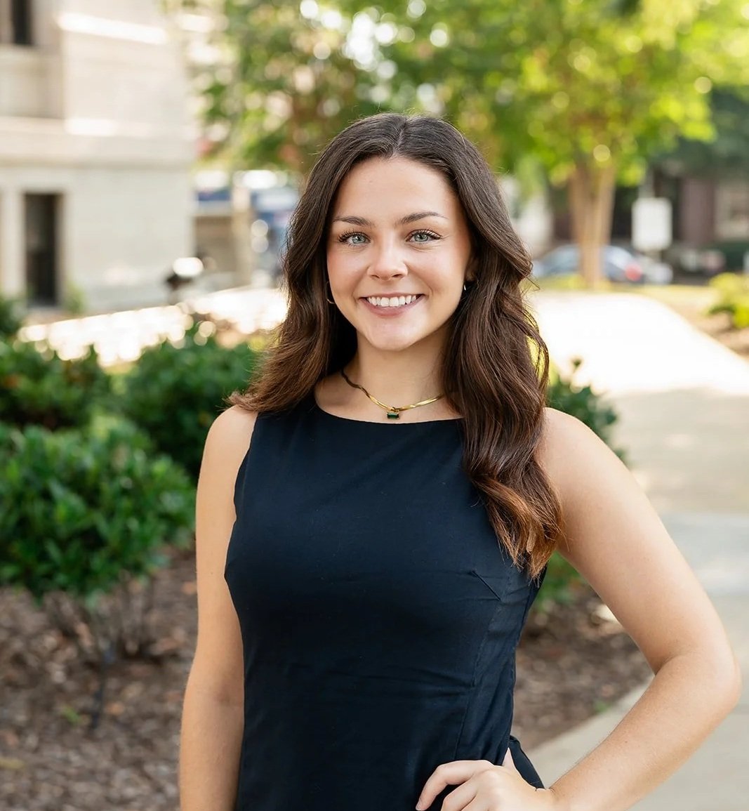 A young woman with long brown hair, wearing a black sleeveless dress and a necklace, standing outdoors with green bushes and trees in the background.