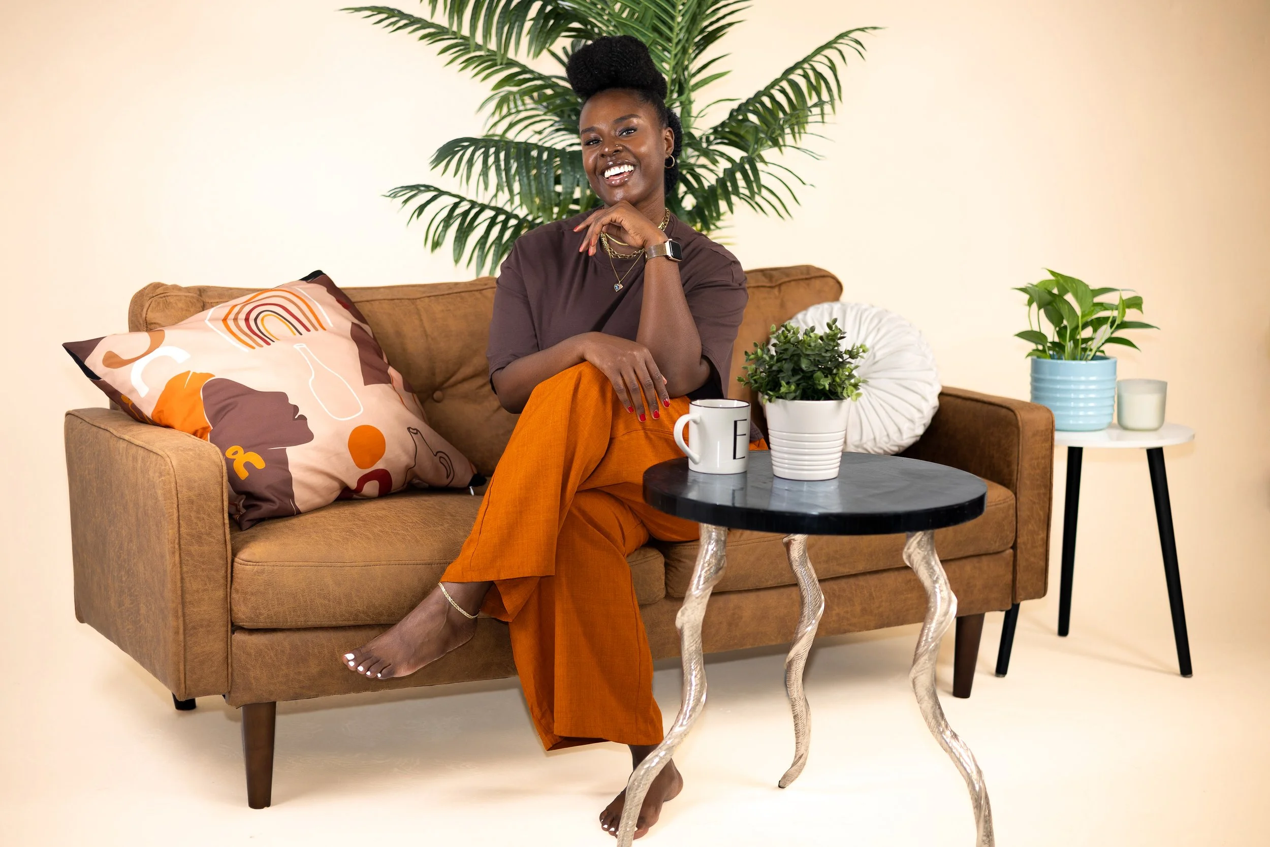 Smiling Black woman in orange pants sitting on a brown couch in a cozy living room with plants and coffee table