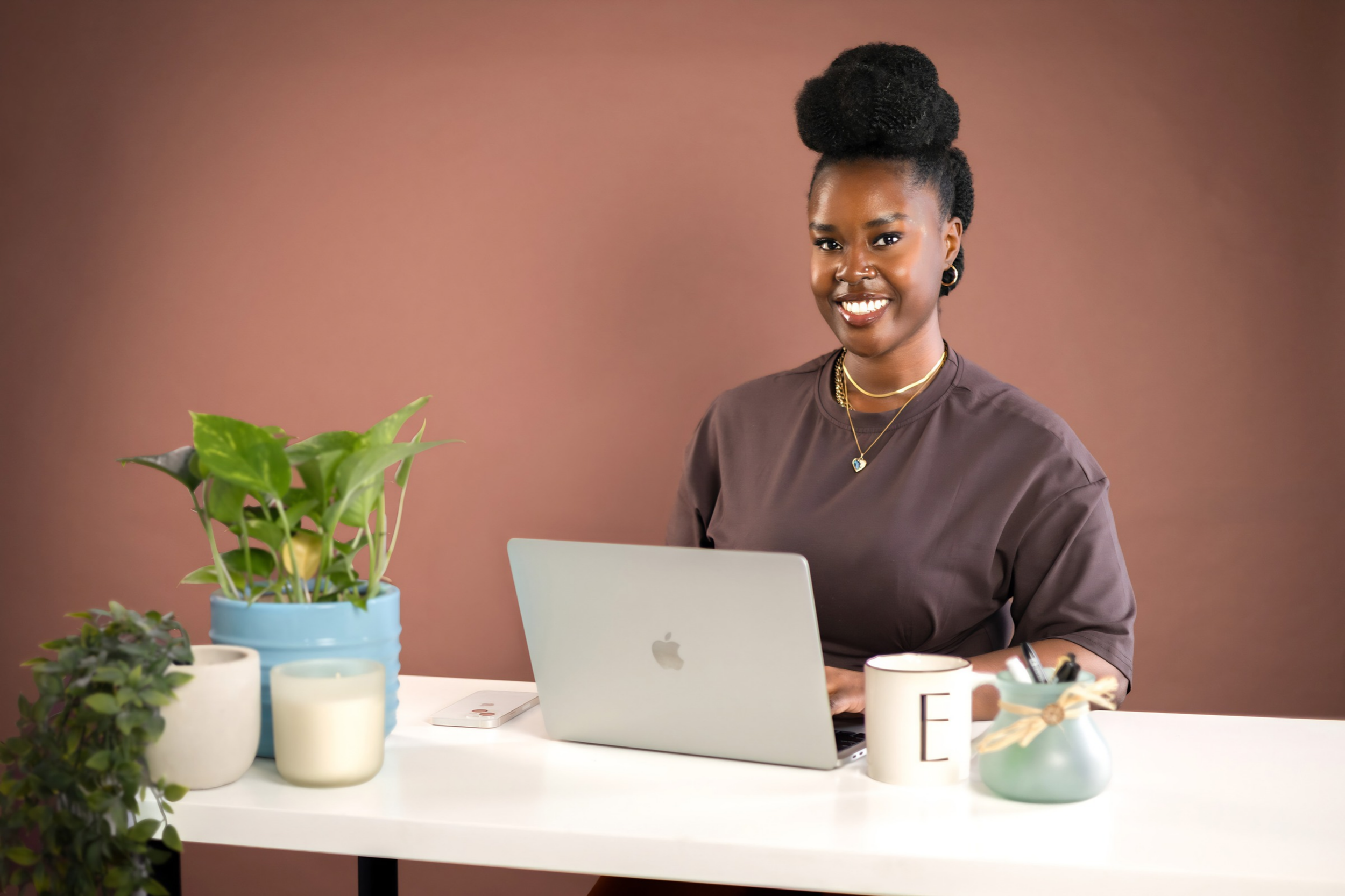 Smiling Black woman working on a laptop at a desk with houseplants, candles, and a mug