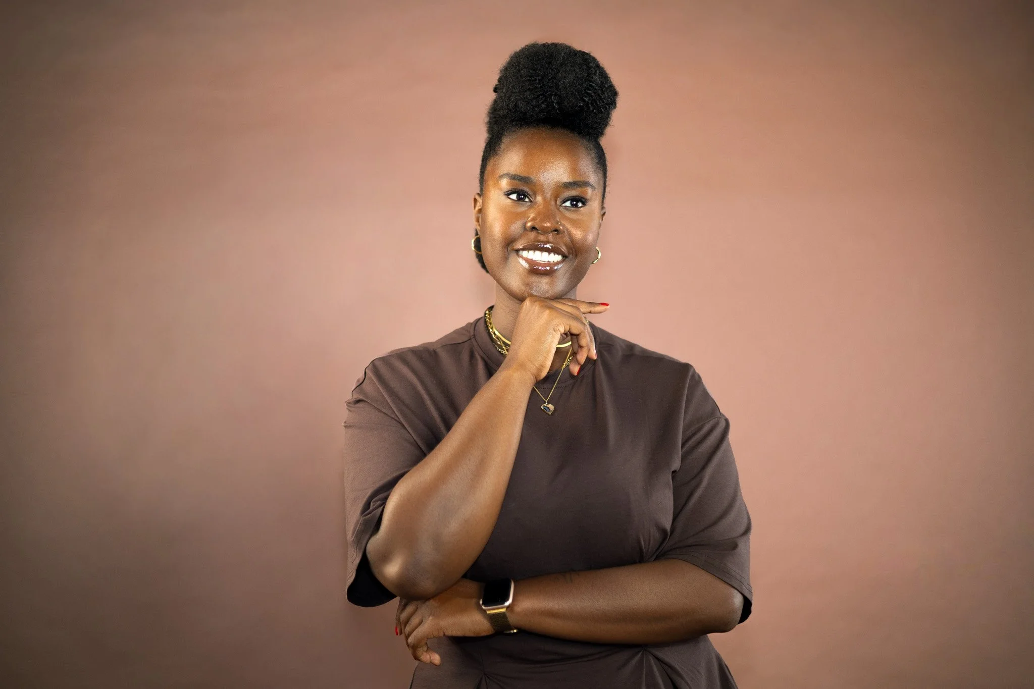 Smiling Black woman with natural hair in a high bun, jewelry, and dark shirt posing against a warm pink background
