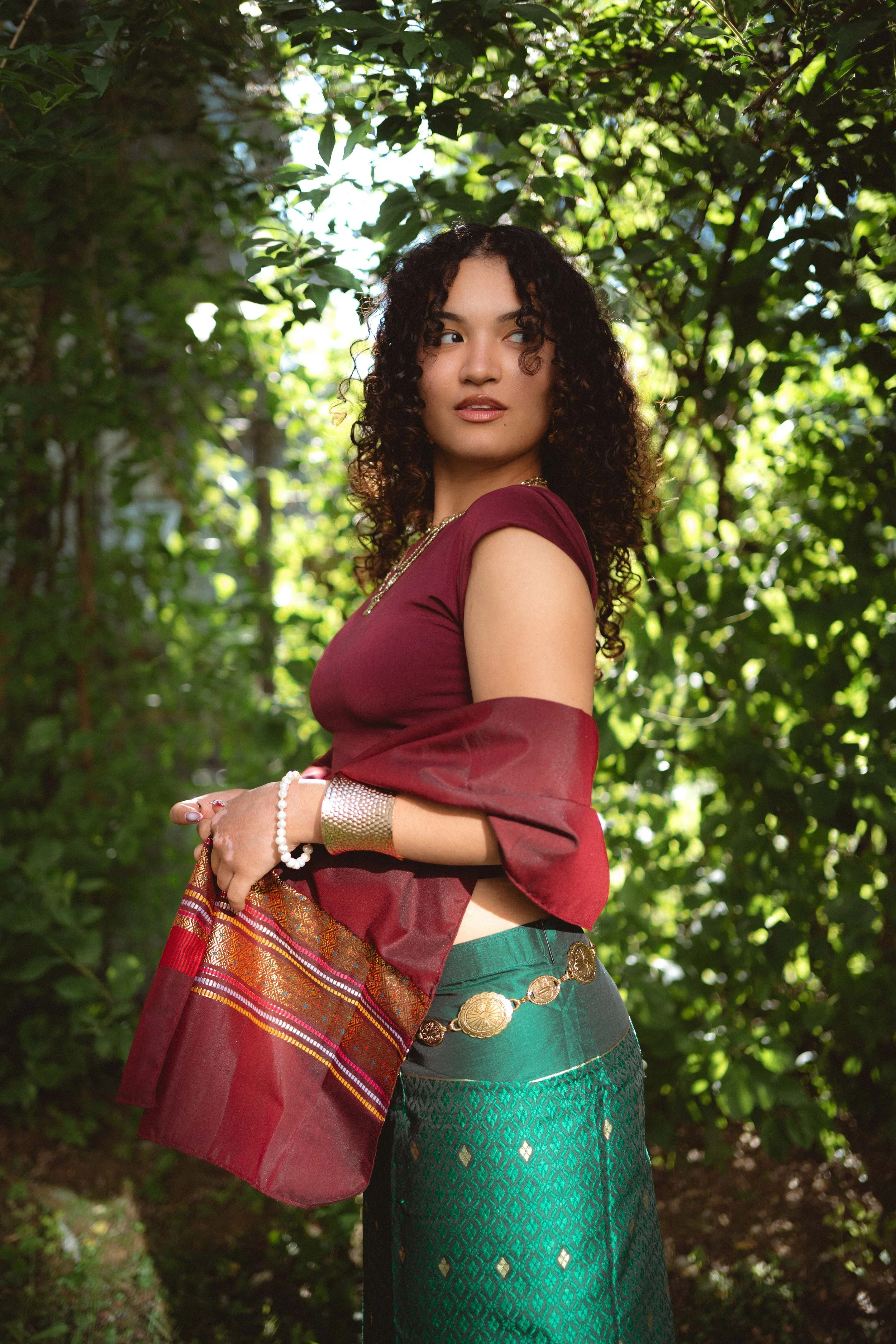 Eine junge Frau in traditioneller philippinischer Tracht mit Grün- und Rottönen, knielang, steht in einem von Bäumen umgebenen grünen Garten, schaut zur Seite, hat lockiges Haar, trägt Schmuck und hat eine elegante Haltung.
