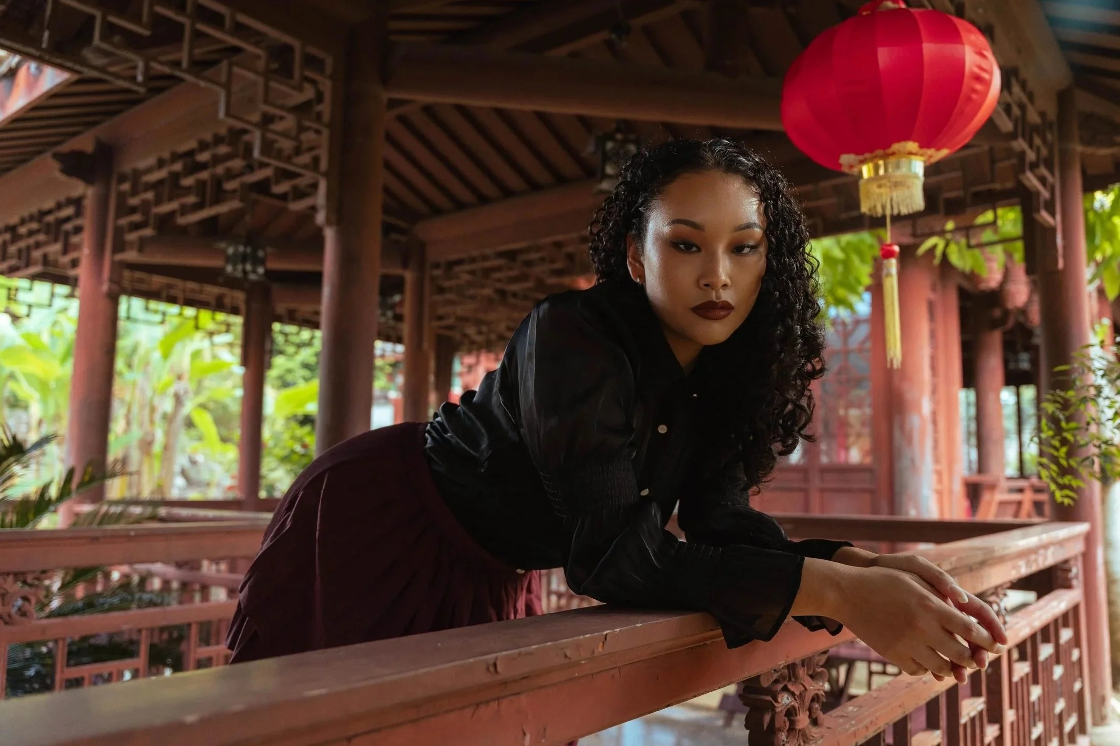 Eine junge Frau mit dunklen, lockigen Haaren und dunklem Lippenstift sitzt an einer roten Brüstung in einer traditionellen asiatischen Umgebung mit Holzarchitektur und roten Laternen.