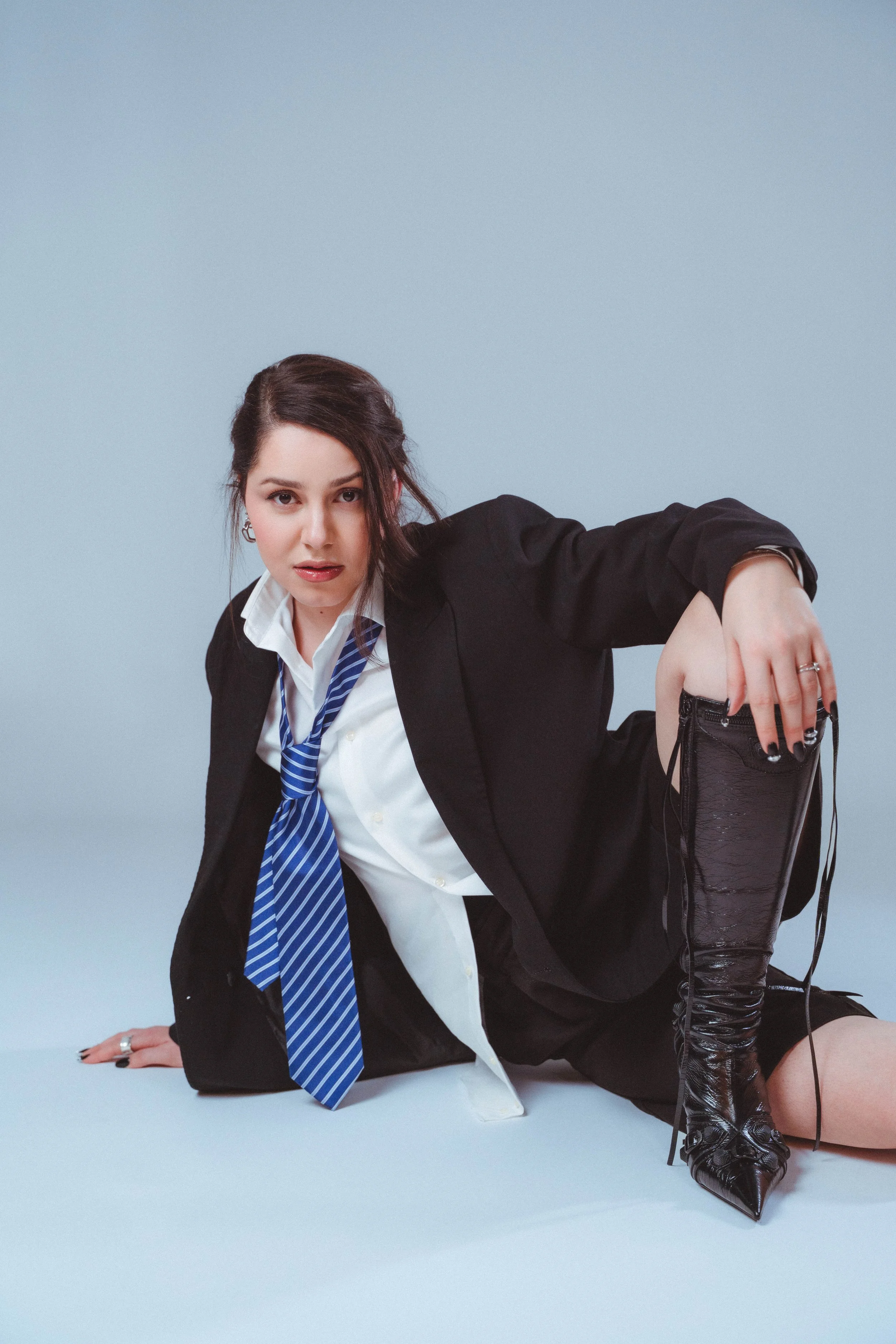 Junge Frau in Business-Outfit, die auf dem Boden kniet, trägt schwarze Stiefel, in der Hand eine Kamera, lässt die Kamera auf den Boden zeigen gegen einen hellblauen Hintergrund.