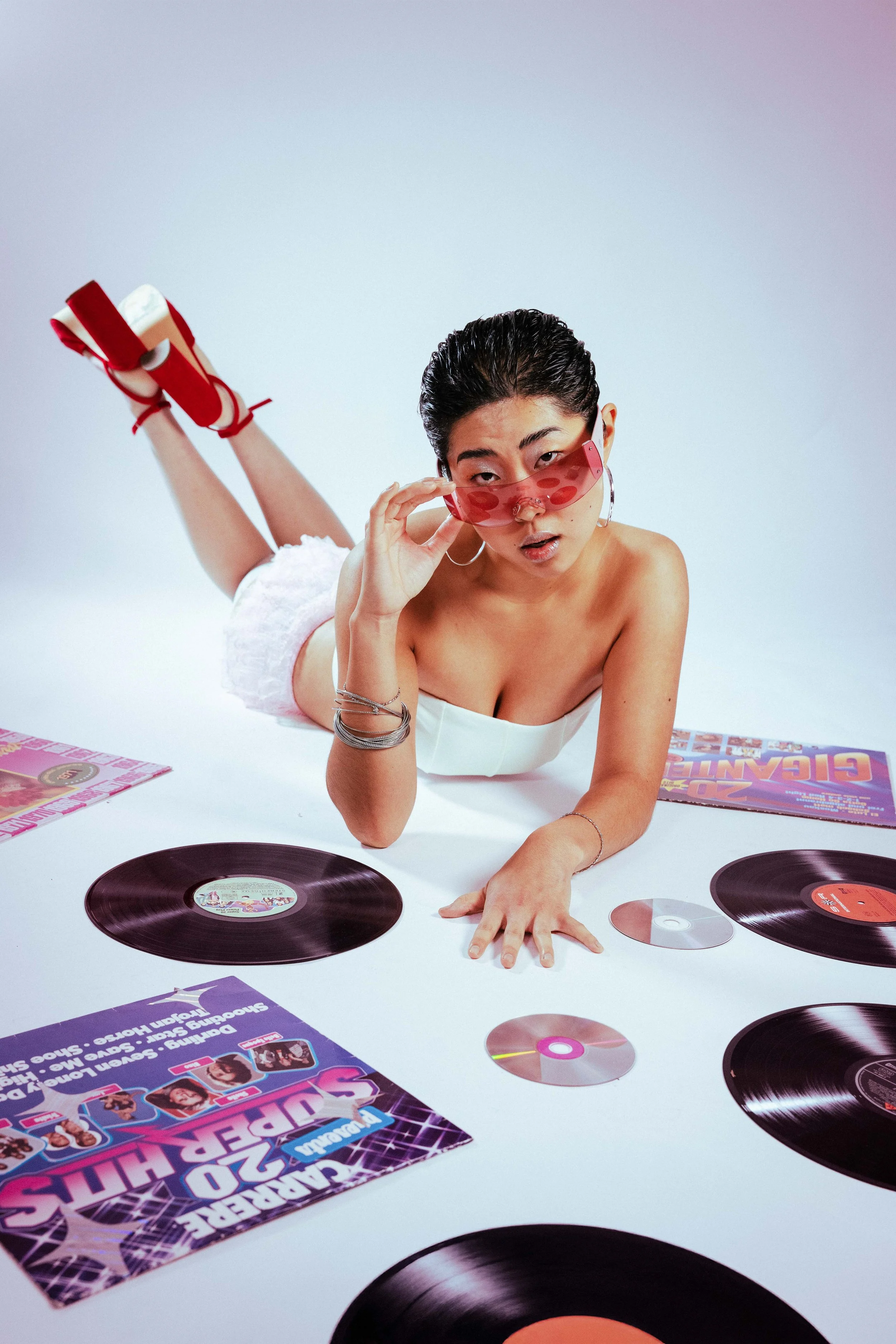 Junge Frau im weißen Top liegt auf dem Bauch, trägt rote Sonnenbrille, hat vor sich Schallplatten, Zeitschriften und Poster, posiert vor weißem Hintergrund.