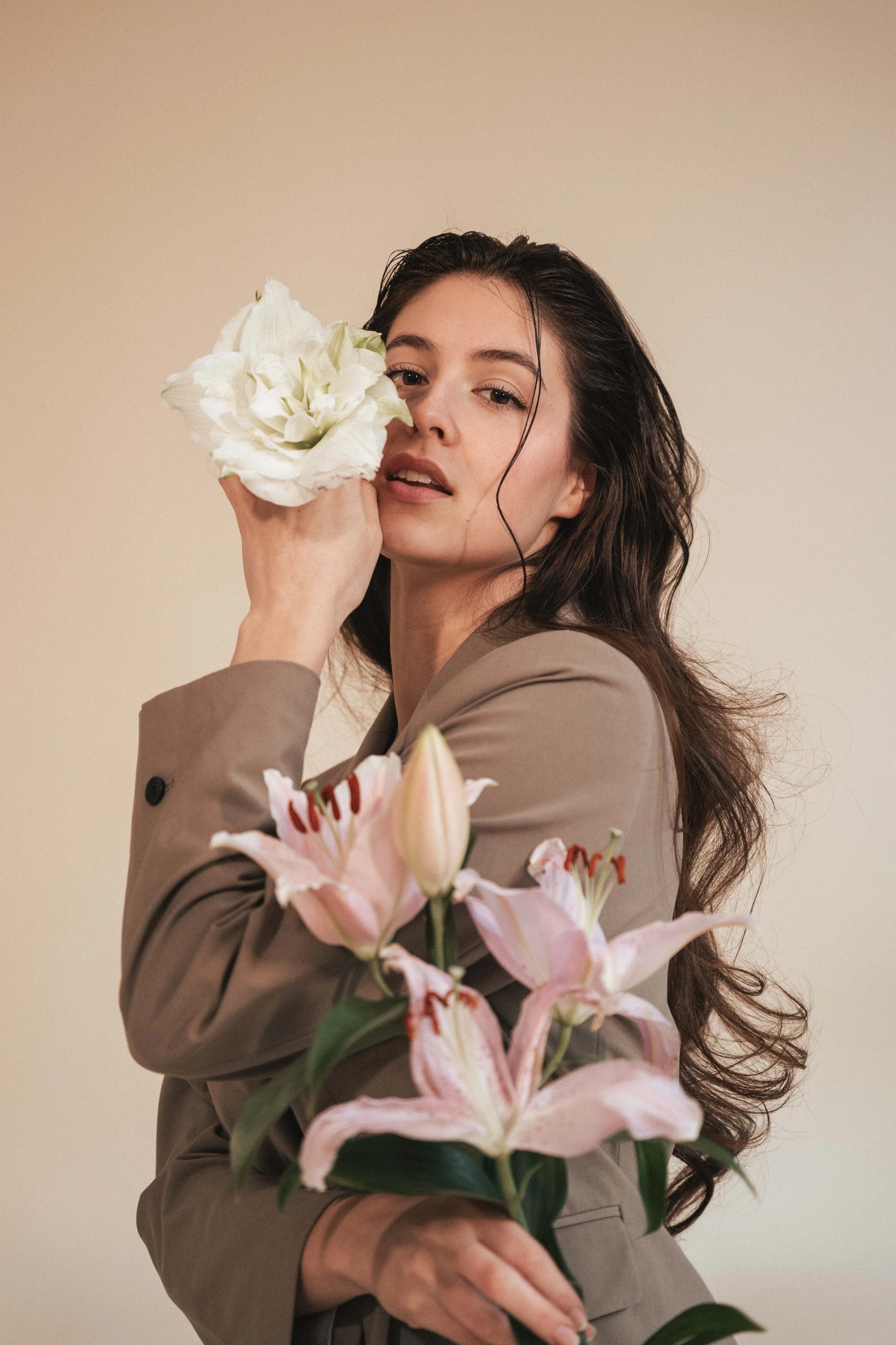 Junge Frau mit langen, dunklen Haaren hält einen weißen Blumenstrauß vor das Gesicht. Im Vordergrund sind pinkfarbene Lilien. Der Hintergrund ist beige.