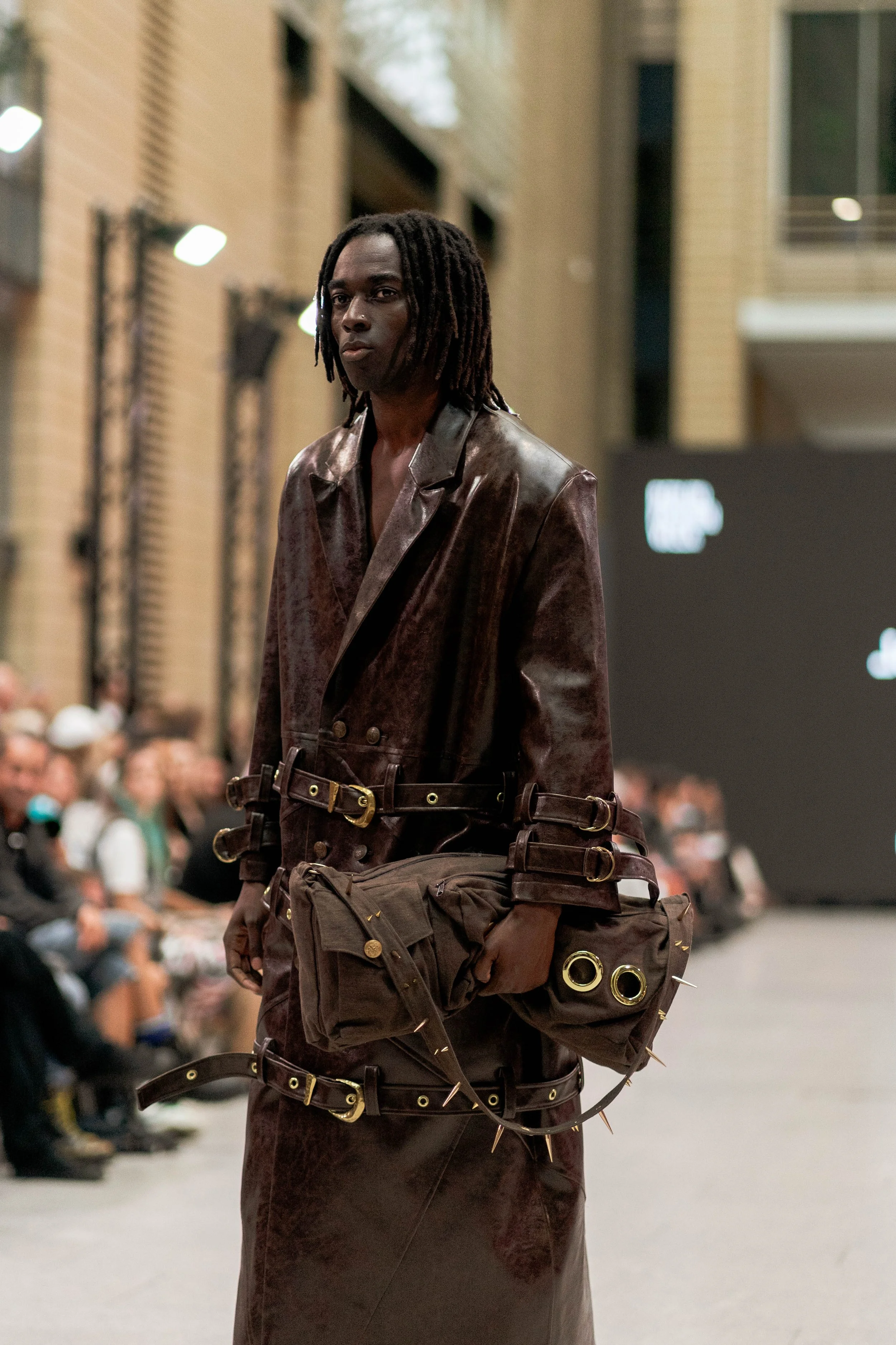 Ein Mann mit schwarzen locks trägt ein braunes Leder-Outfit und trägt eine große braune Tasche, während er auf einer Modeveranstaltung läuft.