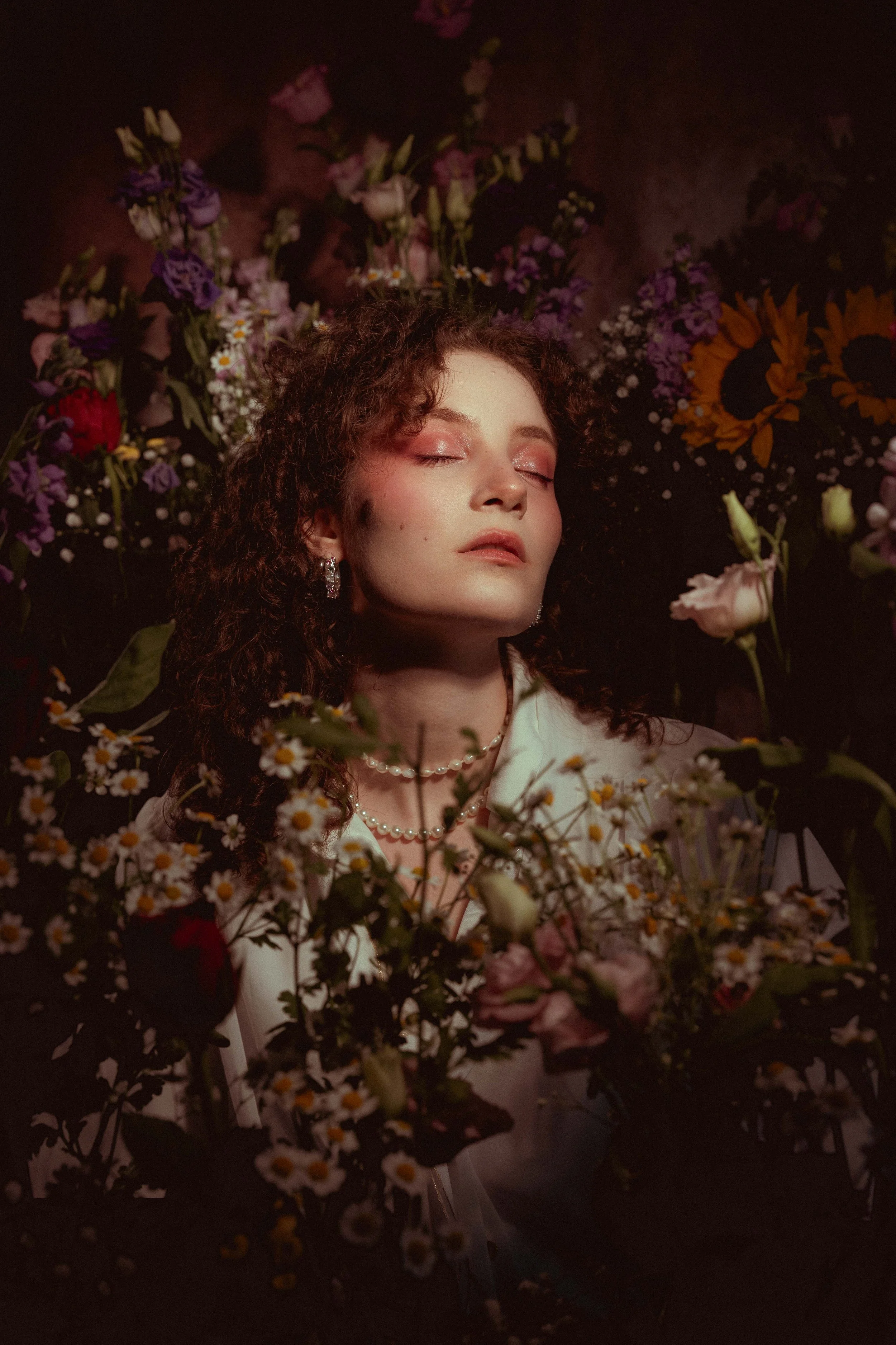 Eine junge Frau mit geschlossenen Augen, umgeben von vielen Blumen, trägt Perlenohrringe und eine Perlenkette, im Hintergrund ist ein dunkler, floraler Hintergrund zu sehen.