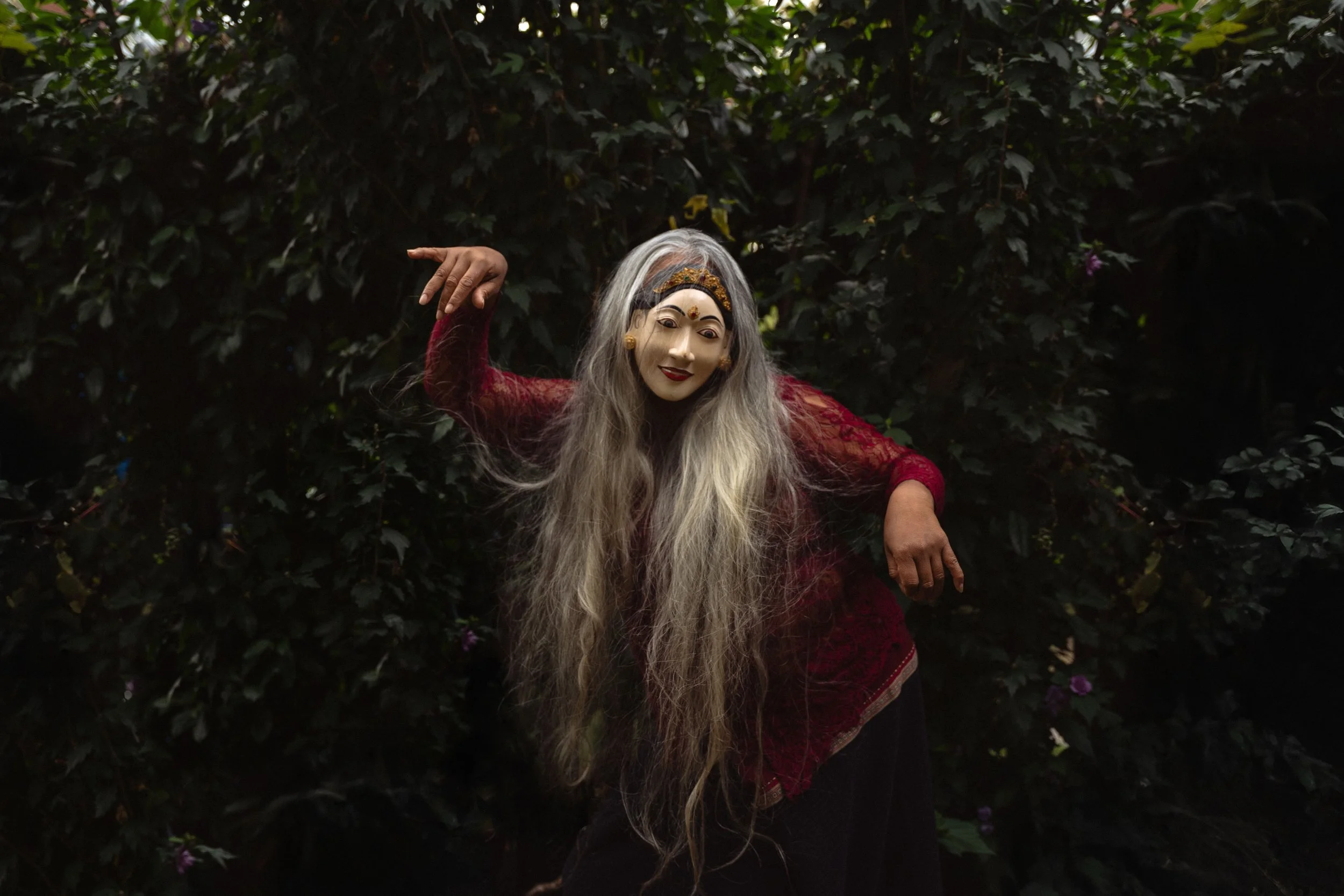 Eine Person trägt eine traditionelle balinesische Maske mit weißen Haaren und einem roten Kleid, posiert vor einem dunklen Laubhintergrund.