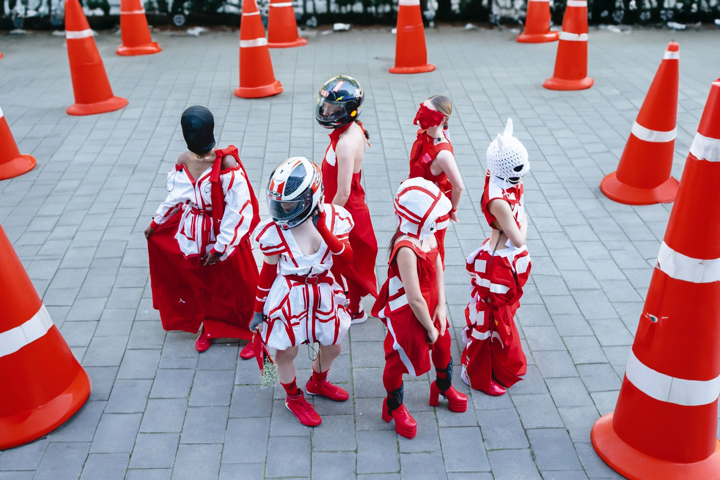 Menschen in roten und weißen Outfits stehen zwischen orangefarbigen Absperrpfosten auf einer gepflasterten Fläche.
