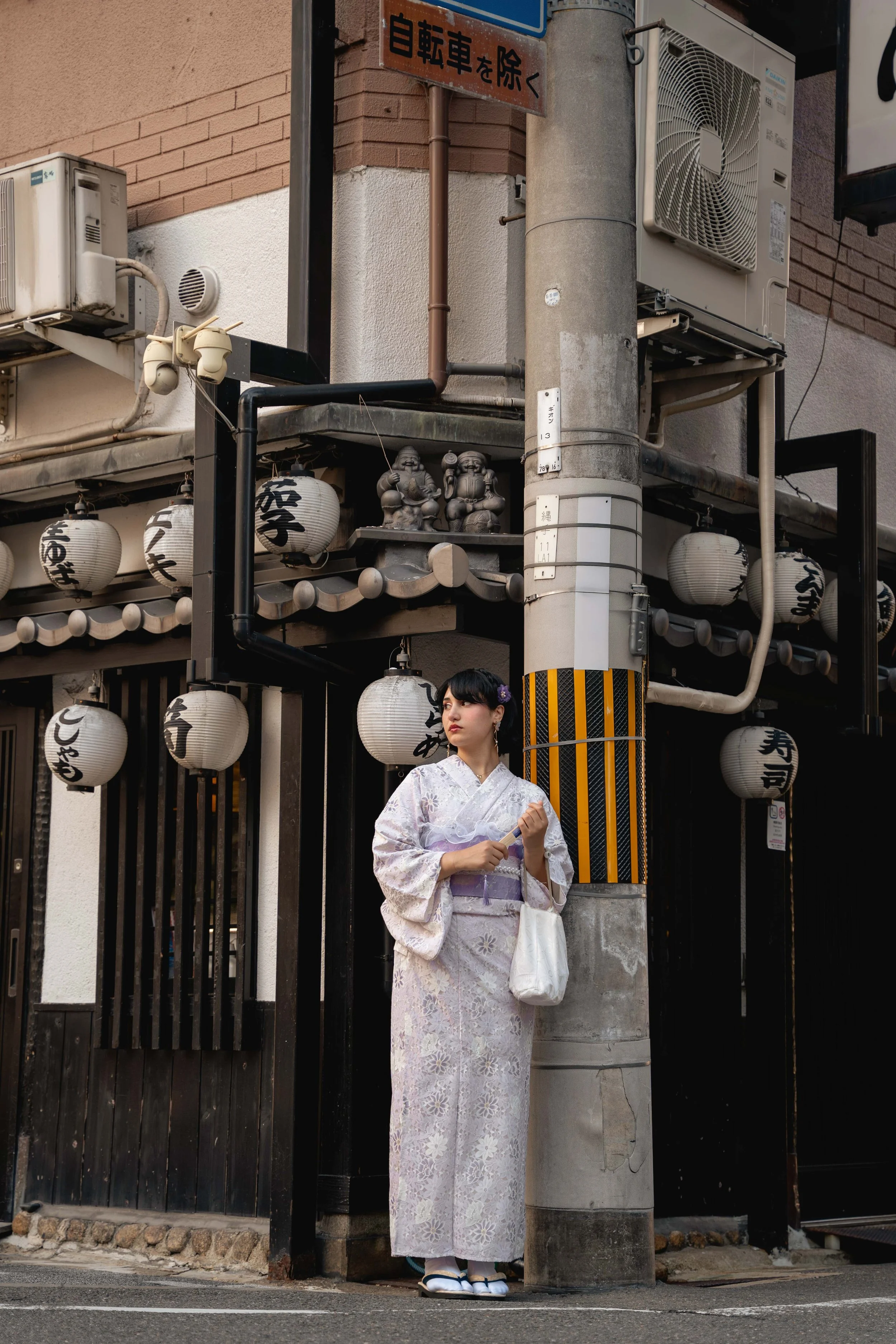 Junge Frau in traditionellem kimono mit weißen Hausschuhen und Handtasche, die auf der Straße vor einem japanischen Restaurant steht, umgeben von Laternen und Stadtmöbeln.