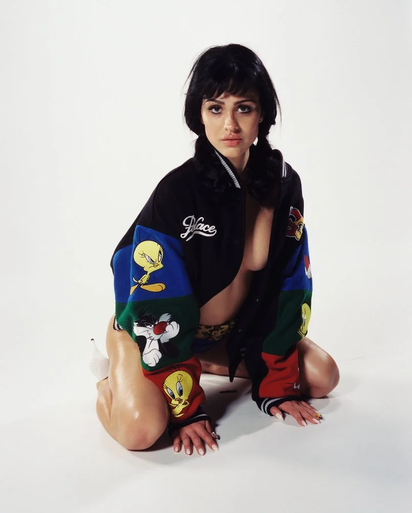 A woman with dark hair and a serious expression is kneeling on the floor with her hands and knees on the ground. She is wearing an oversized black, blue, green, and red jacket with cartoon characters, including Tweety Bird and Sylvester the Cat, on the sleeves. The jacket is partially open, revealing her bare chest. The background is plain white.