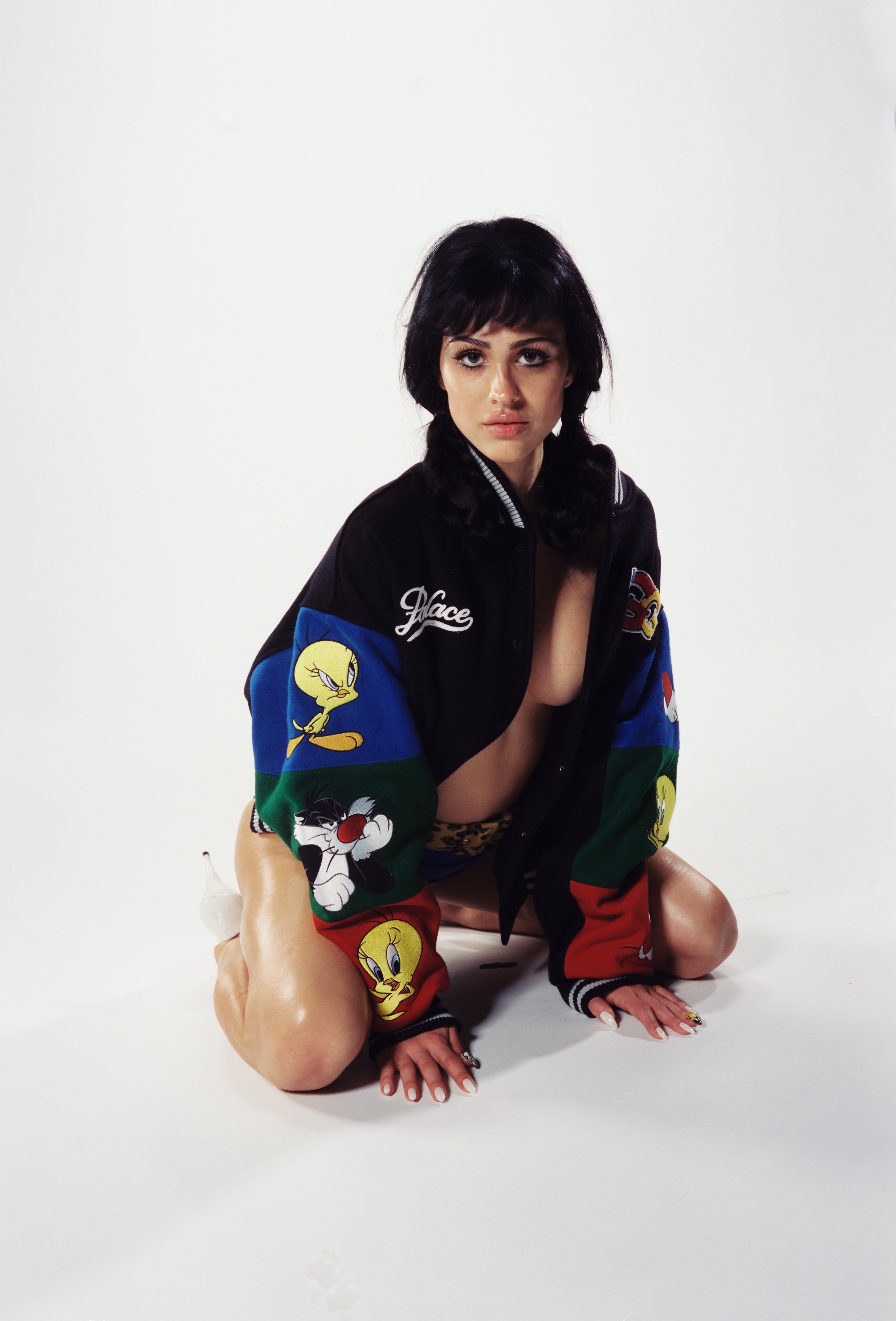 A woman with black hair in pigtails, wearing a colorful cartoon character jacket, is kneeling on the floor in a studio with a white background.
