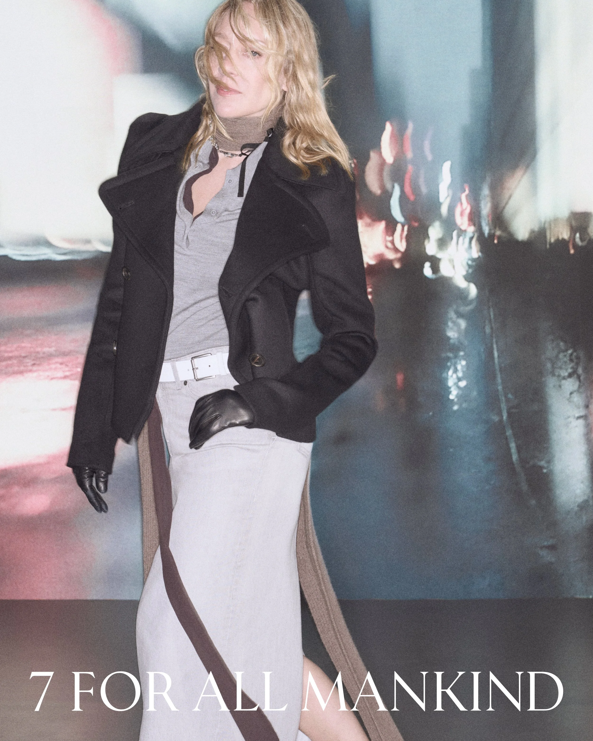 A woman with blonde hair wearing a gray top, black coat, and black gloves standing in front of a cityscape at night with blurred lights.