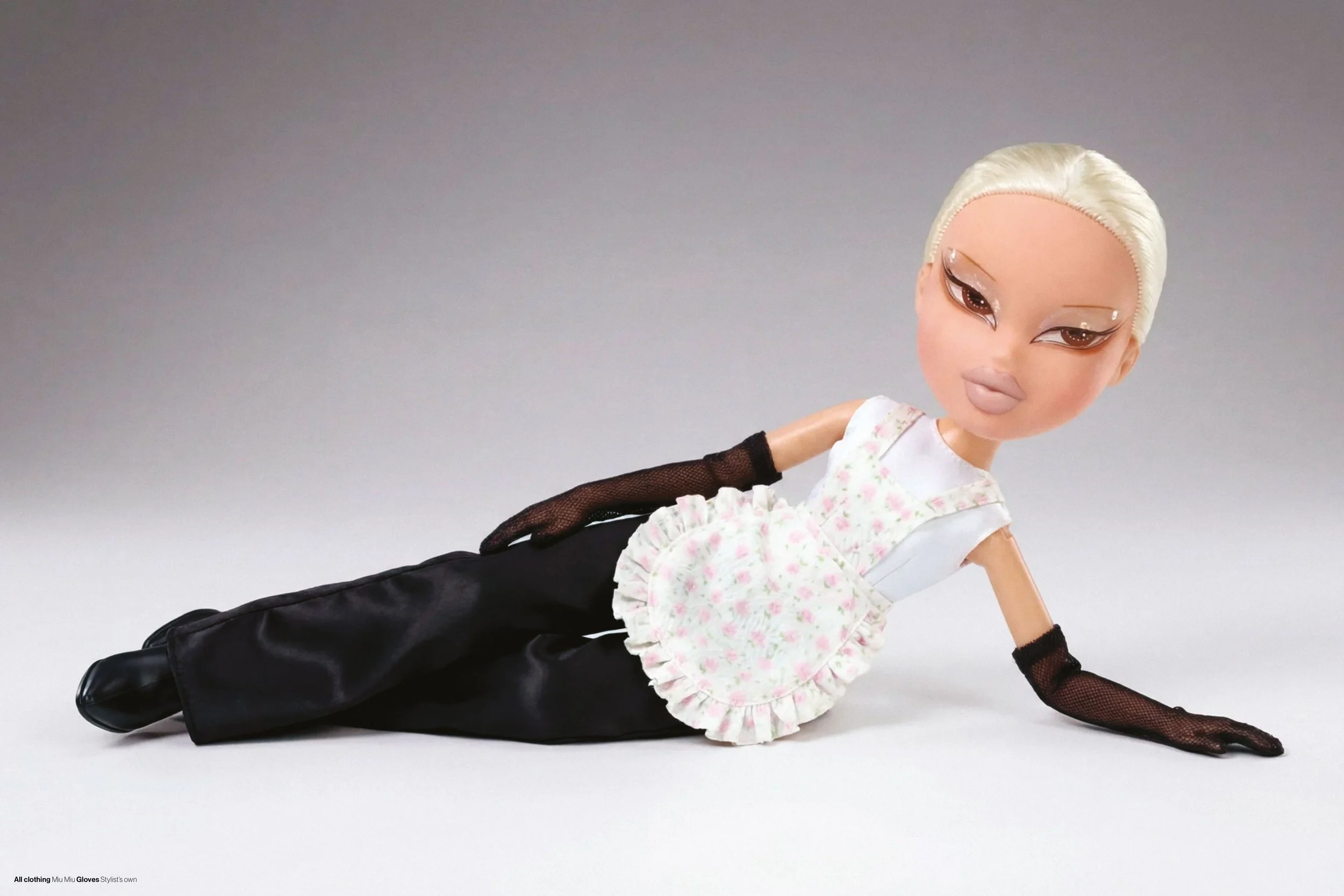 Fashion doll with platinum blonde hair, wearing a floral ruffled top, black satin pants, long black gloves, and black shoes, posed lying on the floor against a neutral background.