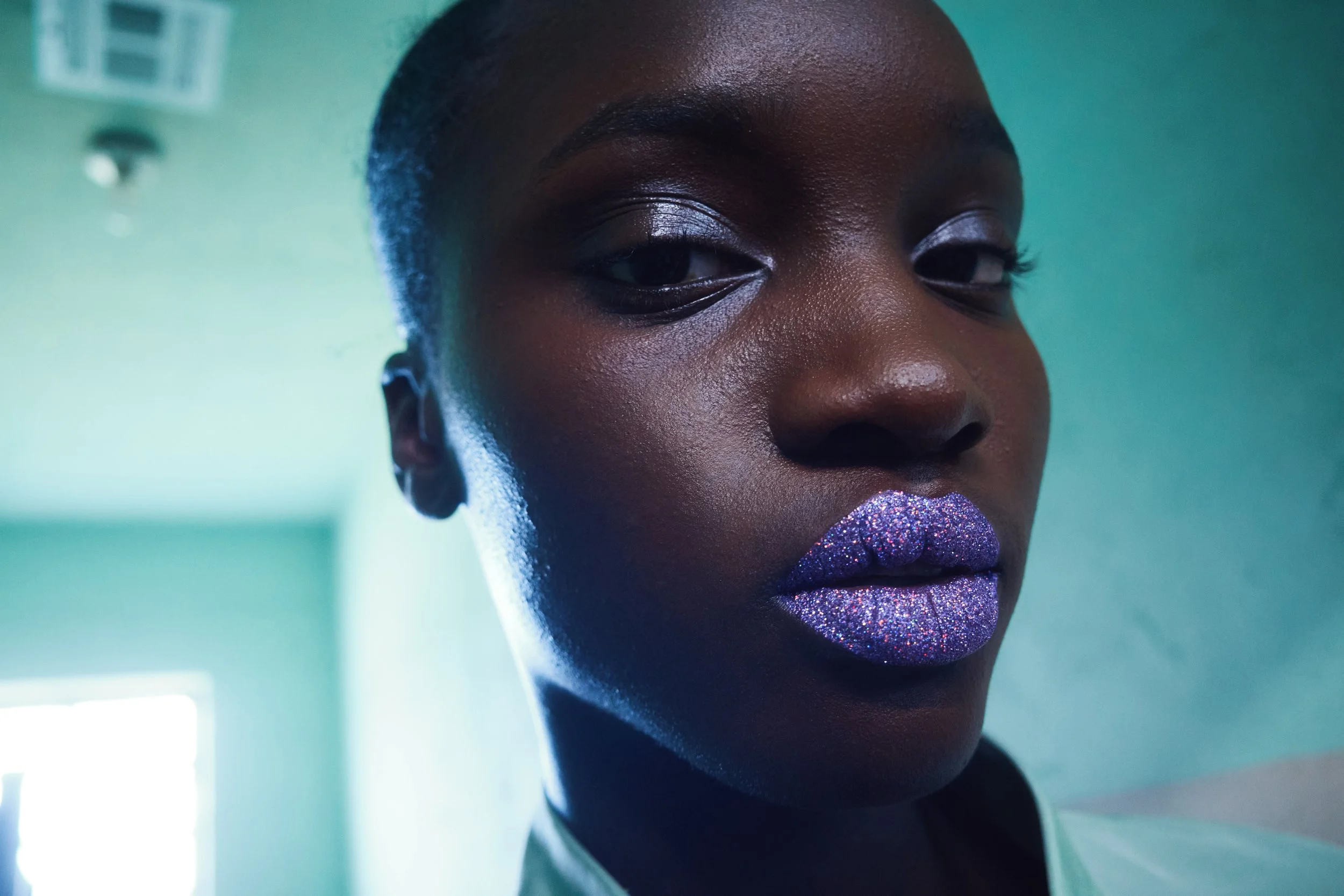 Close-up of a woman with dark skin and shiny glittery purple lipstick, wearing a black top, with a green wall background.