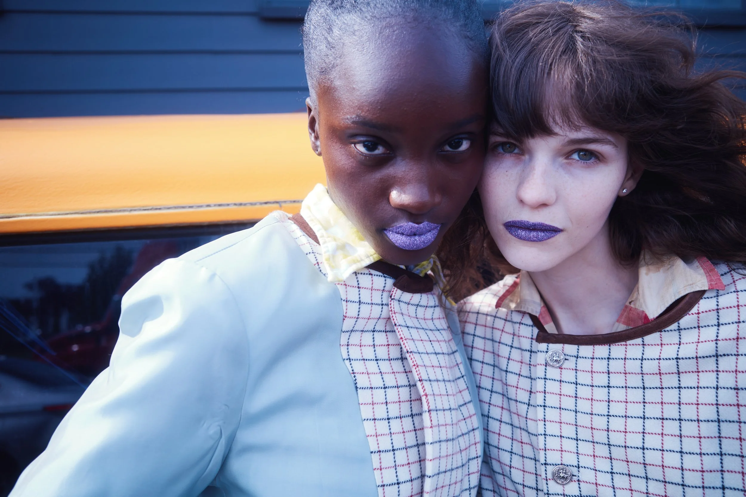Close-up of two women, one with dark skin and the other with light skin, standing close together, both with purple lipstick, in front of a dark wooden background.
