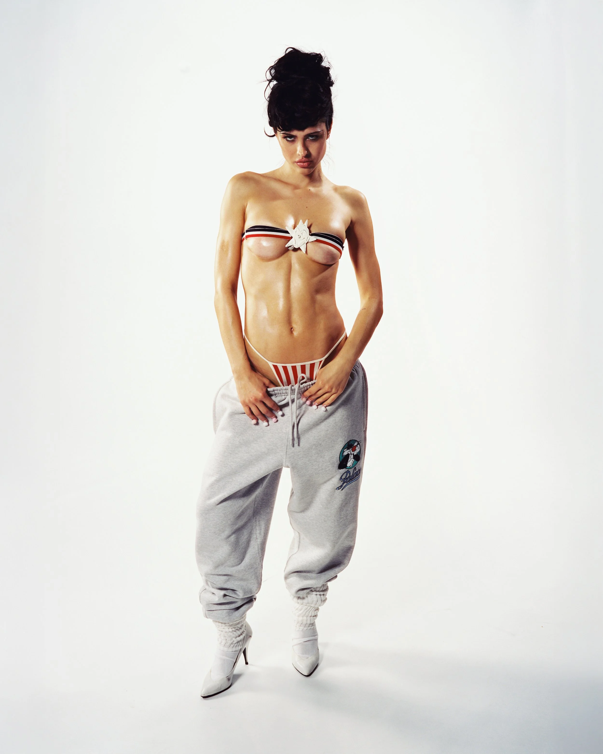 A woman with dark hair styled in an updo, standing against a plain white background. She is wearing white high-heeled boots, gray sweatpants with a graphic on the left leg, and a black, white, and red striped bandeau top with a decorative white bow i