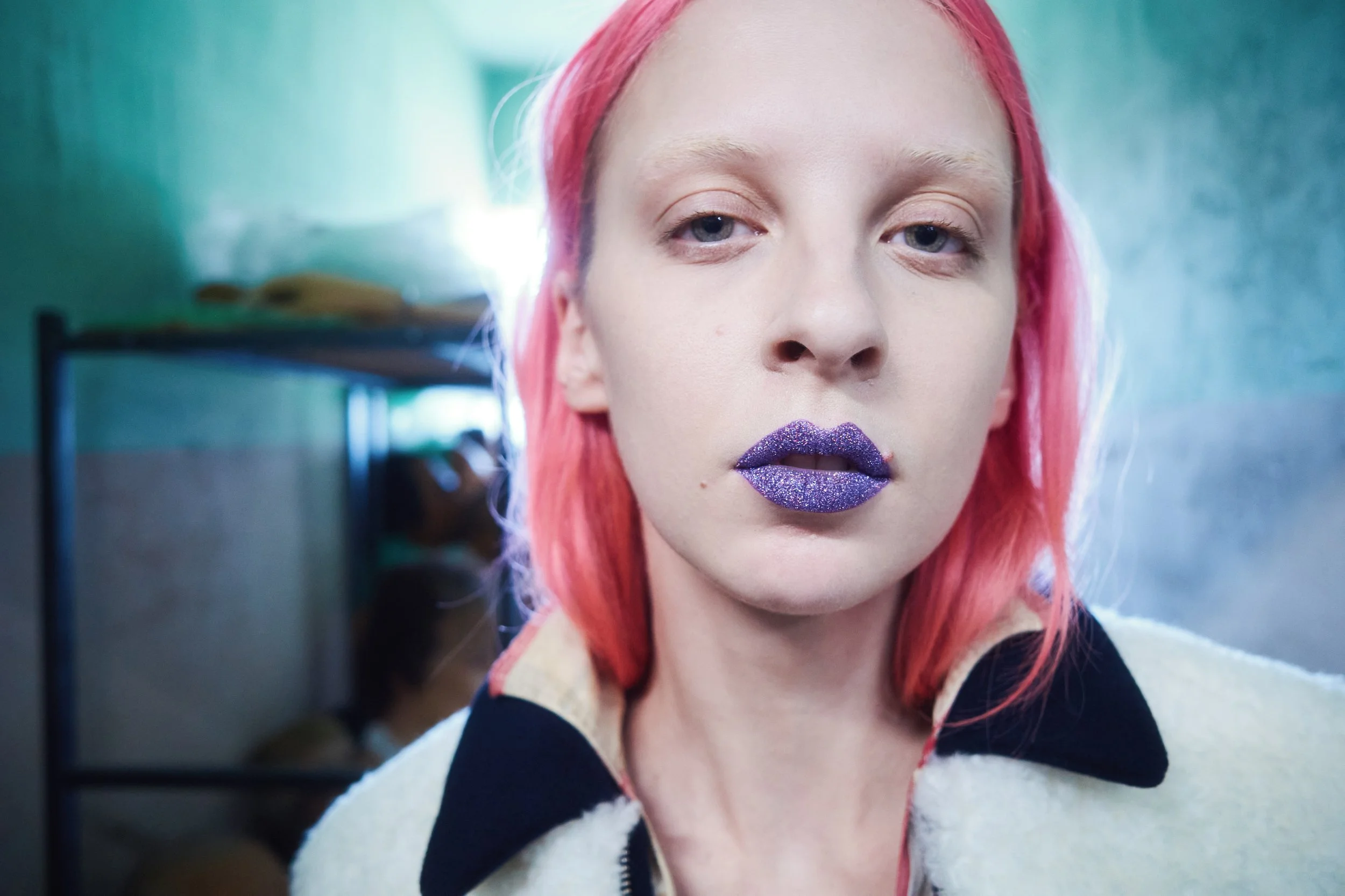 Close-up of a young woman with pink hair and glittery purple lipstick, looking into the camera.