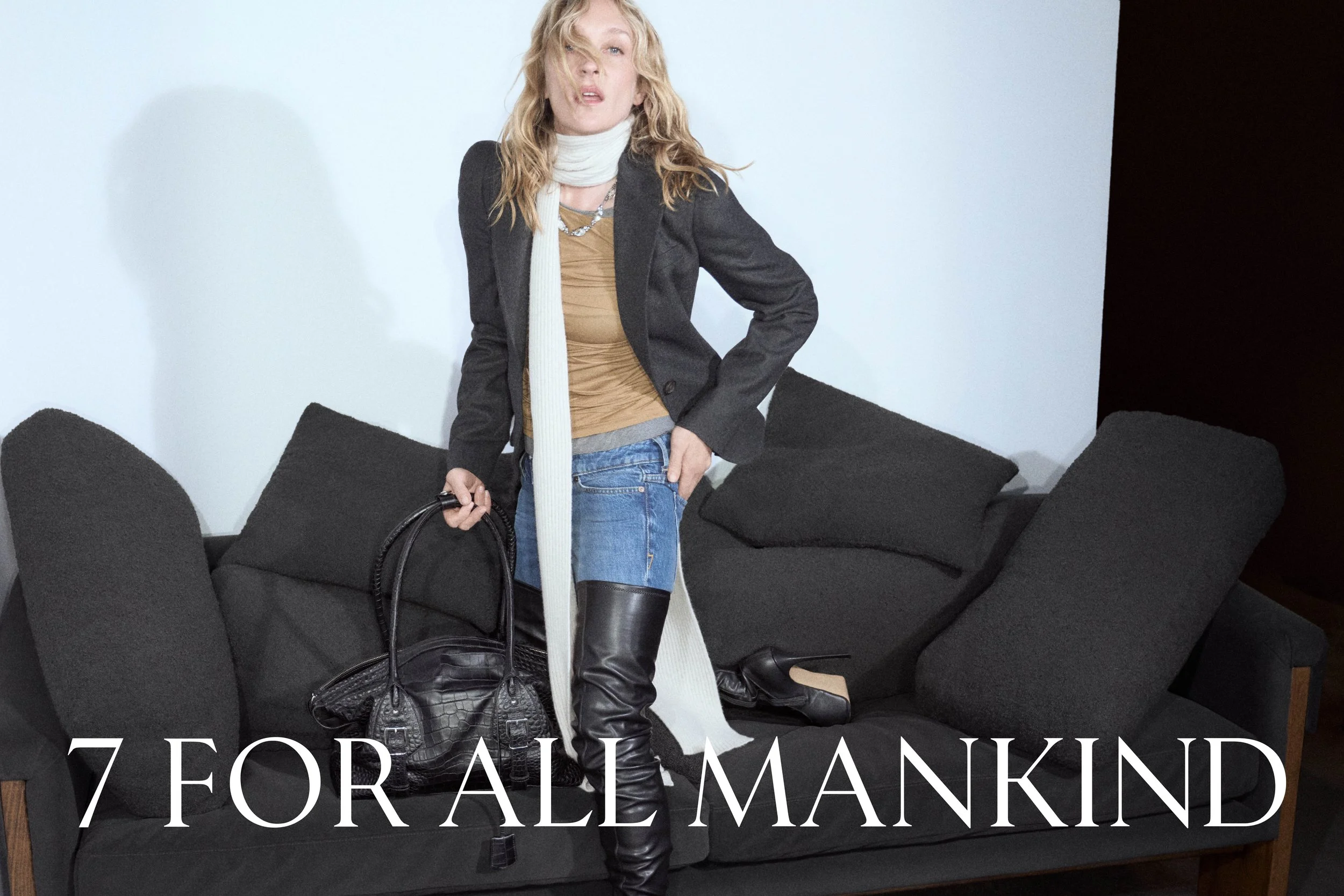 A woman with blonde hair wearing a black blazer, tan top, blue jeans, and black knee-high boots, holding a black handbag, standing in front of a black sofa with pillows. The background is a plain wall with part of a dark wall on the right side. White