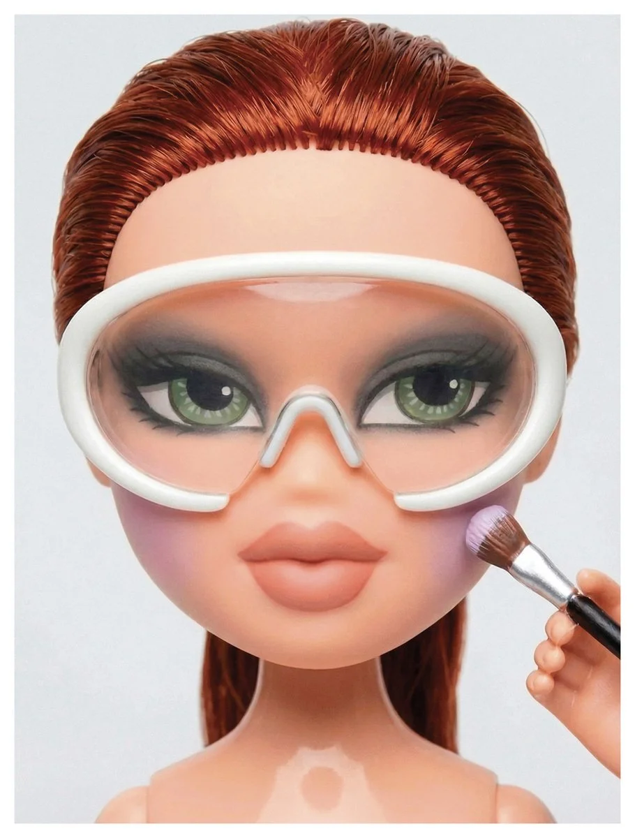 A doll with green eyes, red hair, and pink lips wearing goggles is having makeup applied with a small brush.