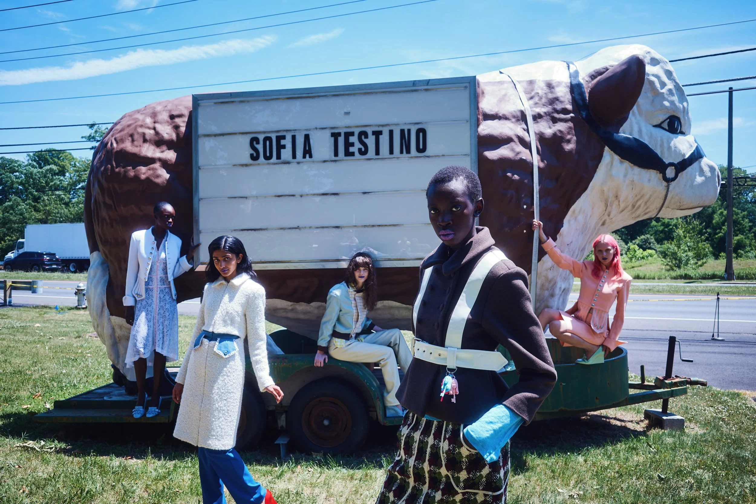 People dressed in vintage fashion pose around a parade float shaped like a horse with a sign that reads "Sofia Testino." The float is on a grassy area beside a road with cars visible in the background, under a blue sky with wispy clouds.