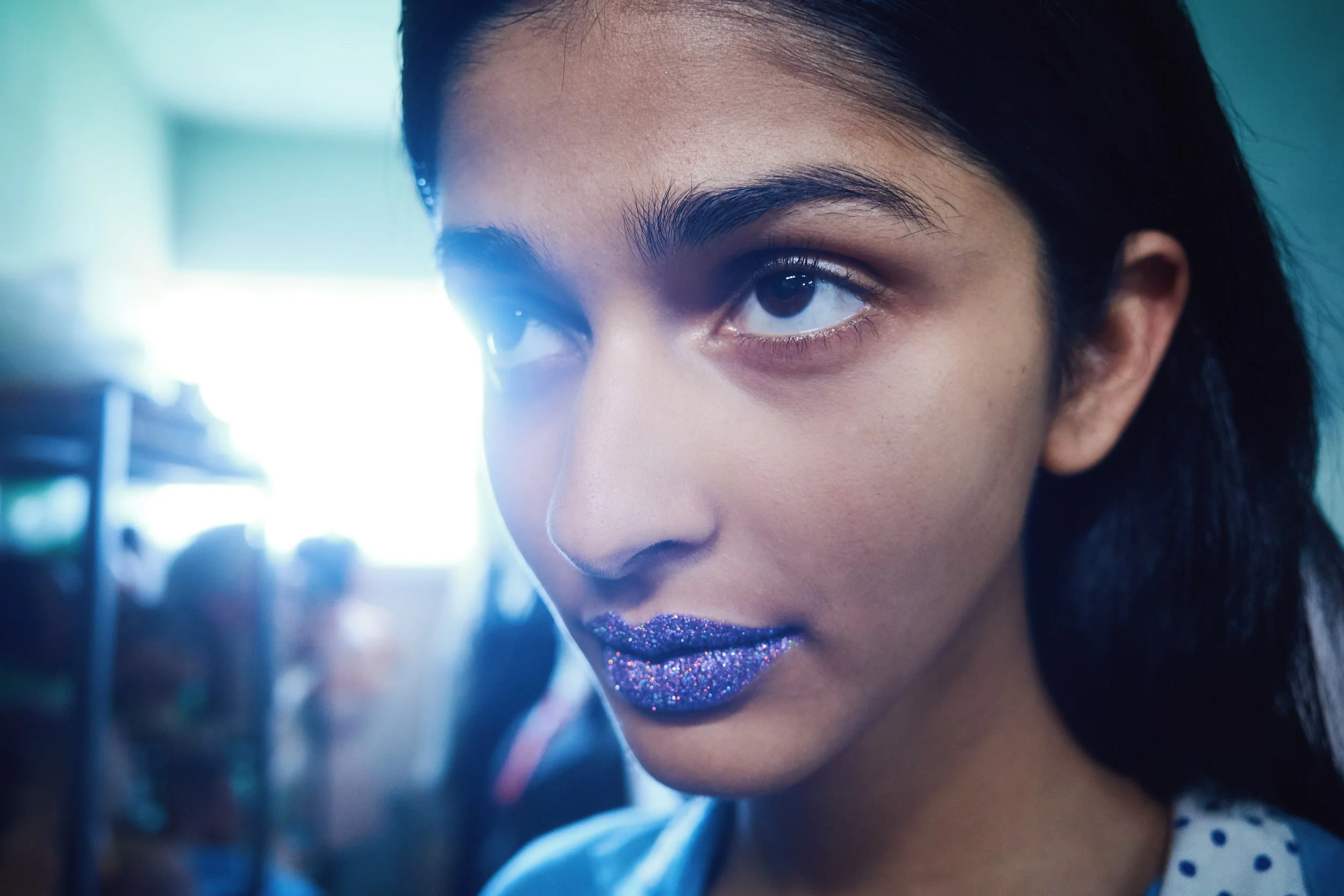 Close-up of a woman's face, focusing on her eye and forehead, with glitter on her lips and dark hair.