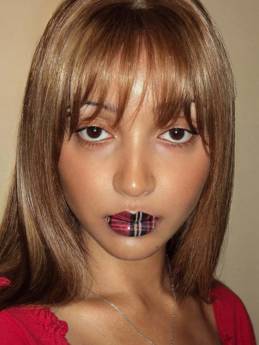 A woman with shoulder-length brown hair with bangs, wearing a red top and a silver necklace, has a close-up facial portrait with makeup, including shiny lipstick with a plaid pattern, and neutral background.