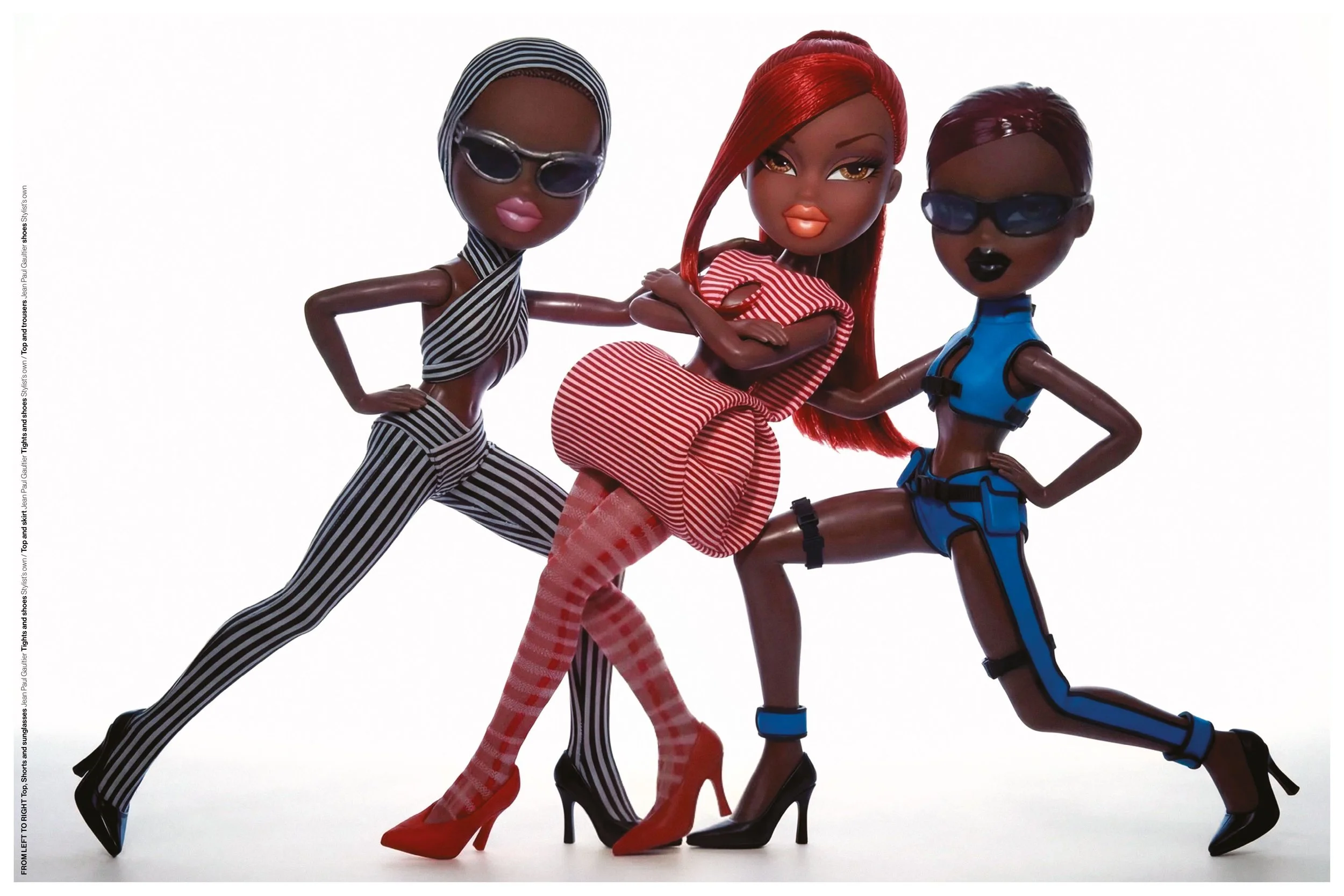 Three fashion dolls with dark skin tones posing together. The doll on the left wears black and white striped top and pants with sunglasses, the middle doll has long red hair, orange lips, red and white striped dress, and high heels, and the doll on t